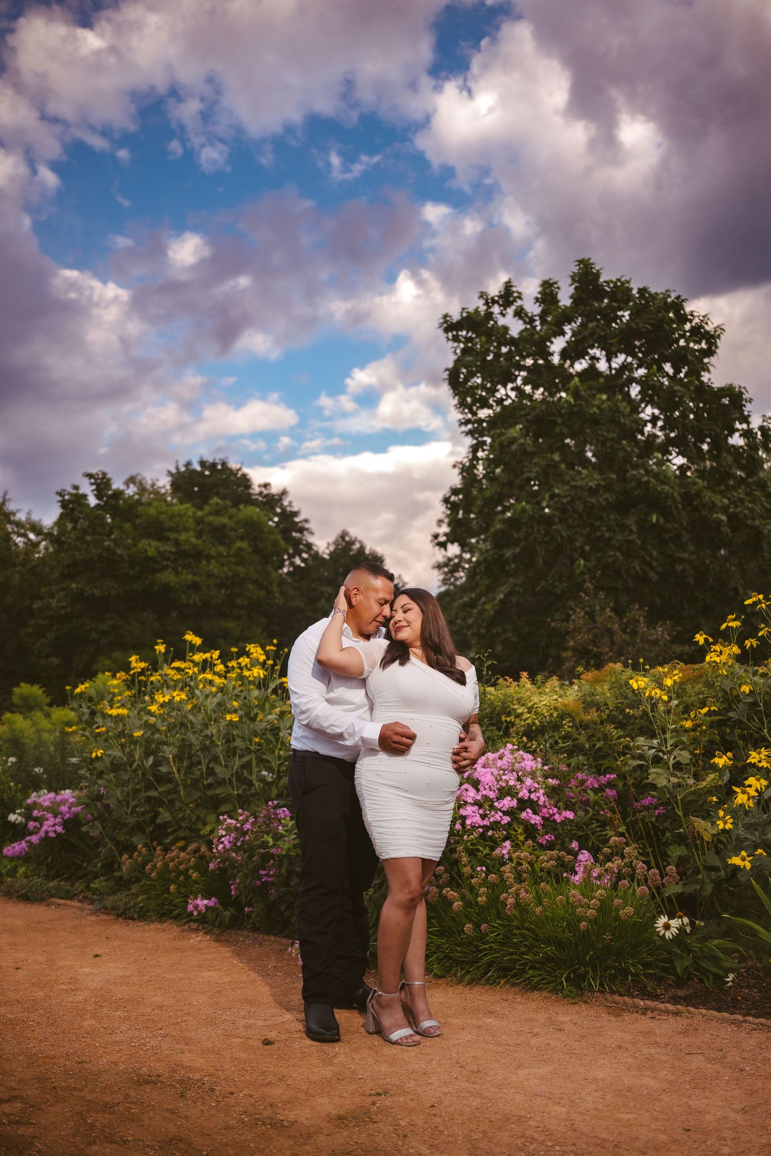 Two people dance and embrace on a garden path with colorful flowers and dramatic sky in the background.