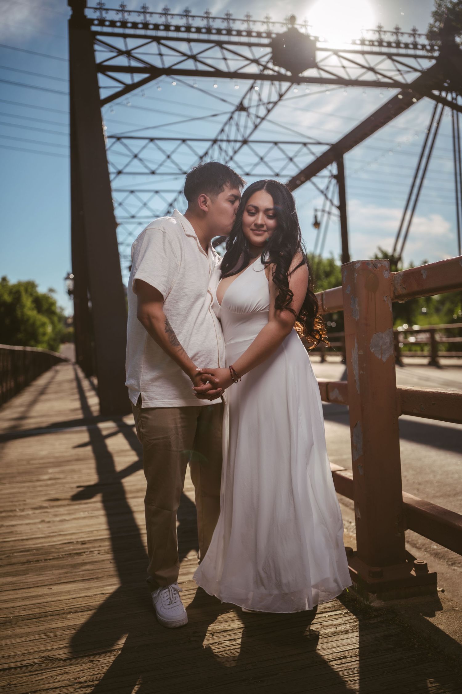 Couple embracing on a vintage iron bridge in warm sunlight, man kissing woman in white maxi dress.