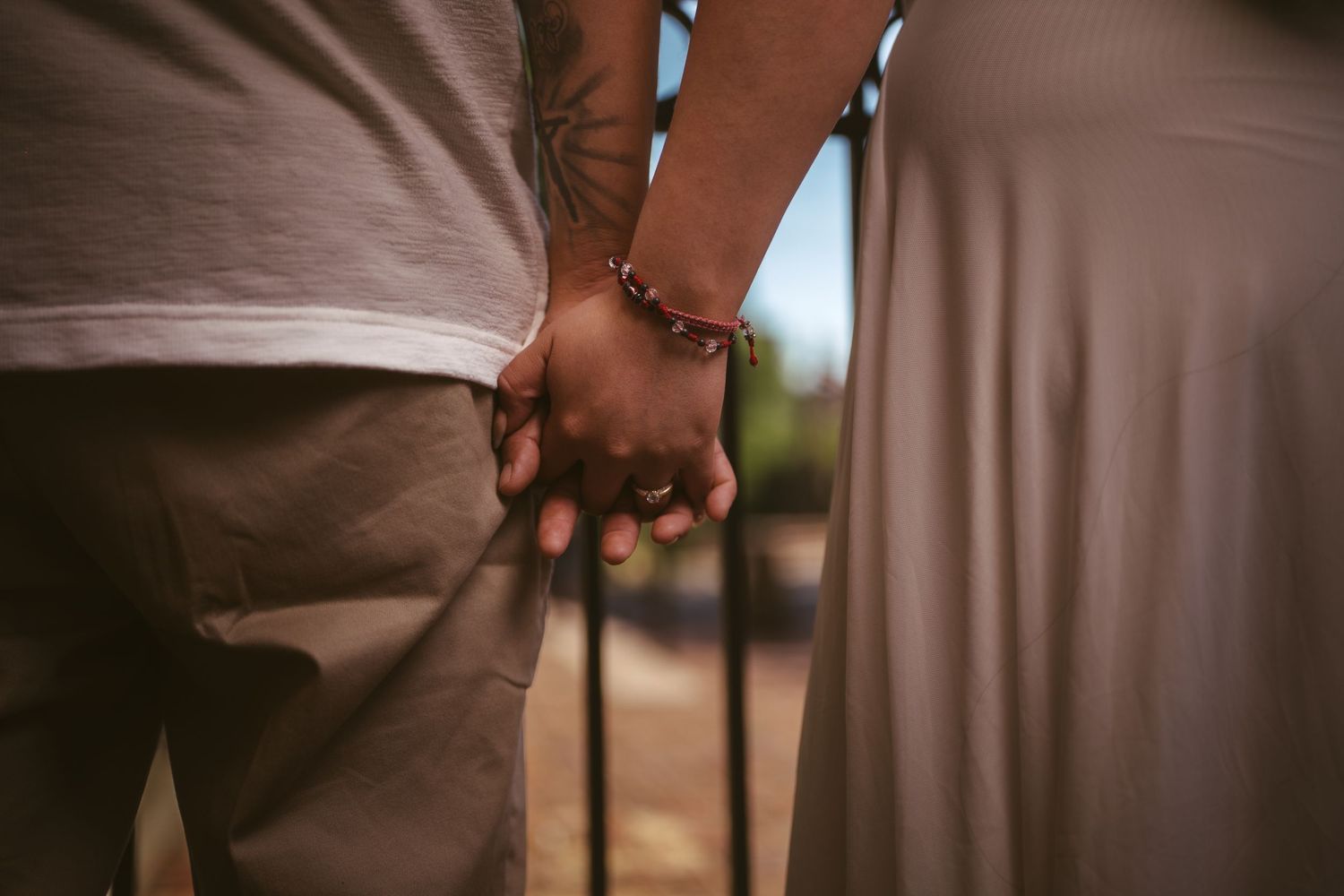 Close-up of a couple holding hands, man wearing a beaded bracelet, standing near iron fence.