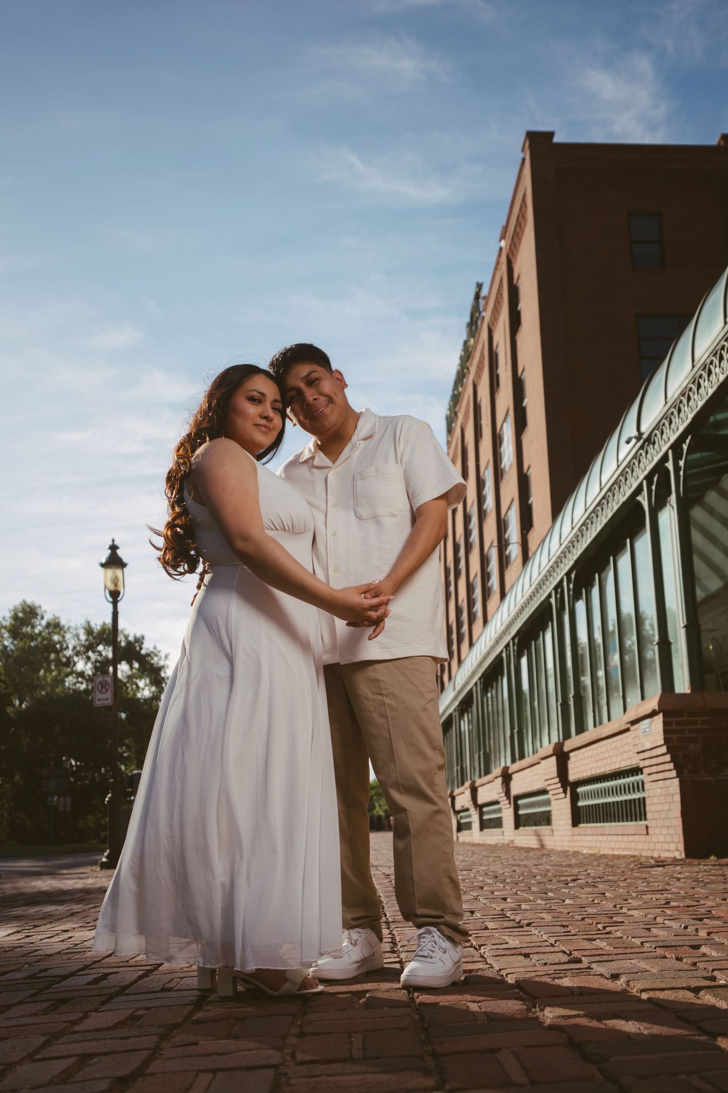 Couple in white dress and khakis hold hands on cobblestone street beside historic brick building at sunset.