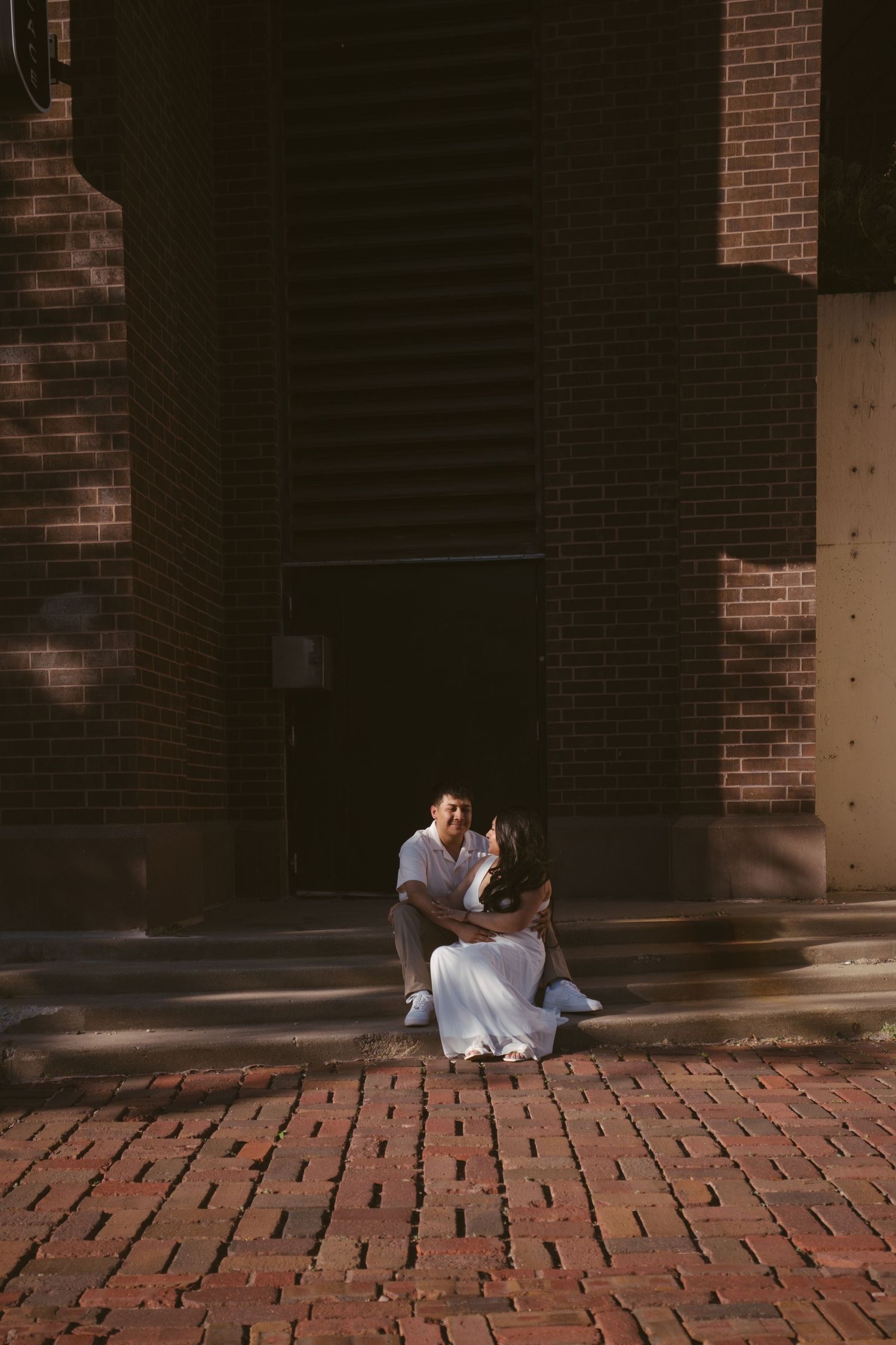 Couple sitting together on brick steps outside a historic red brick building in warm sunlight.