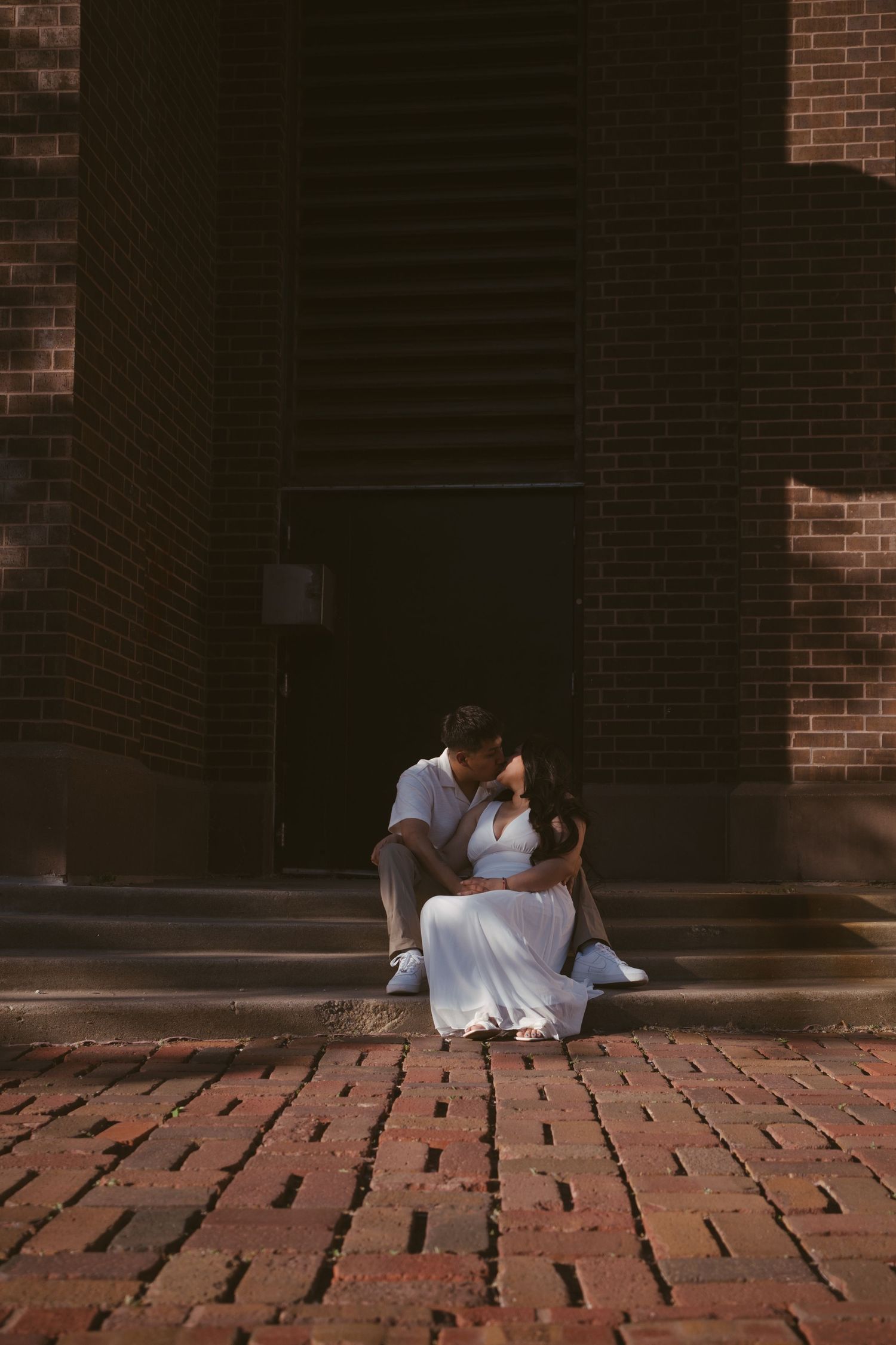 Couple sitting on brick steps sharing a romantic kiss in front of a dark doorway of a brick building.