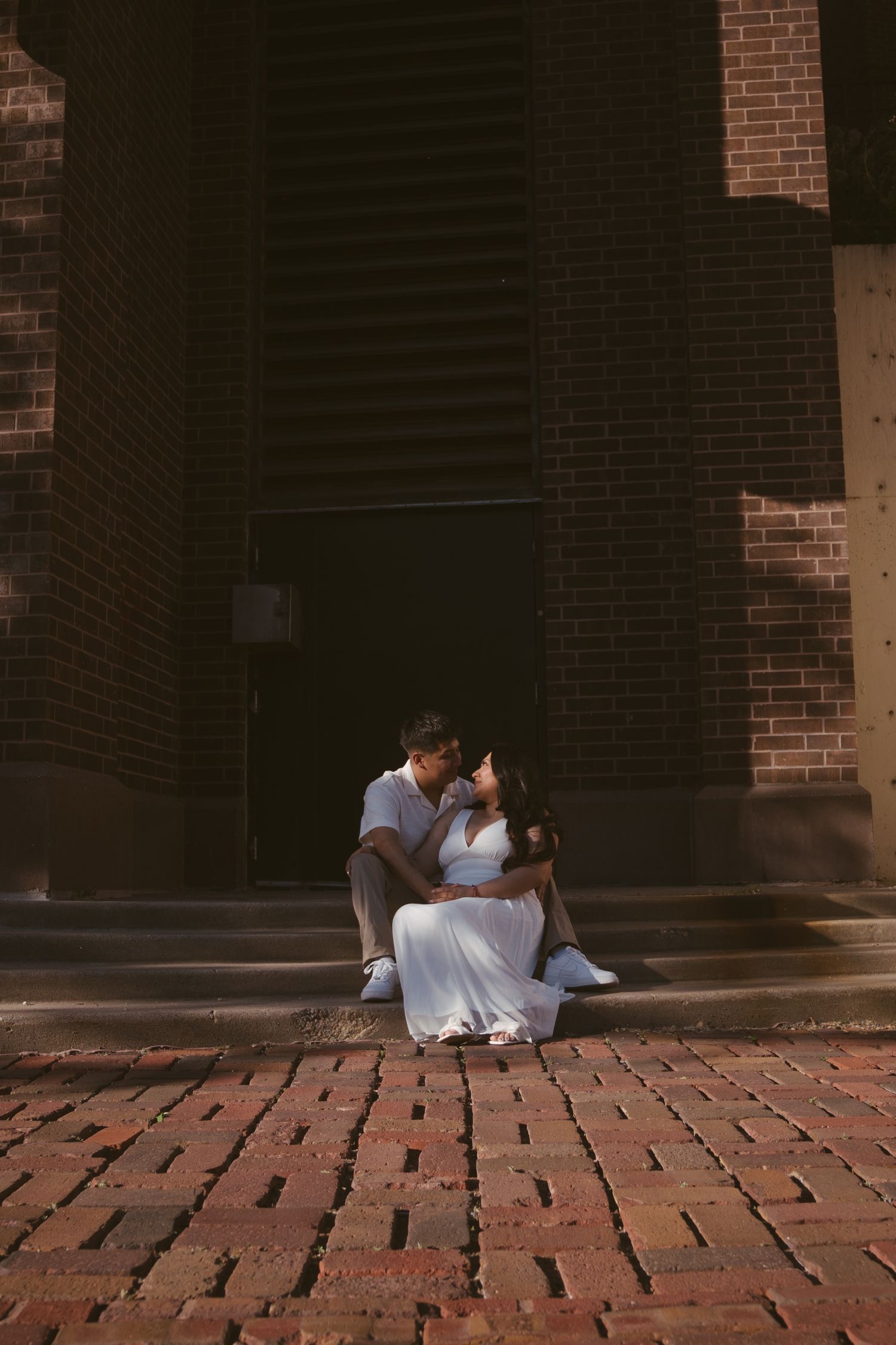 Romantic couple sharing a kiss while sitting on brick steps outside a historic building.