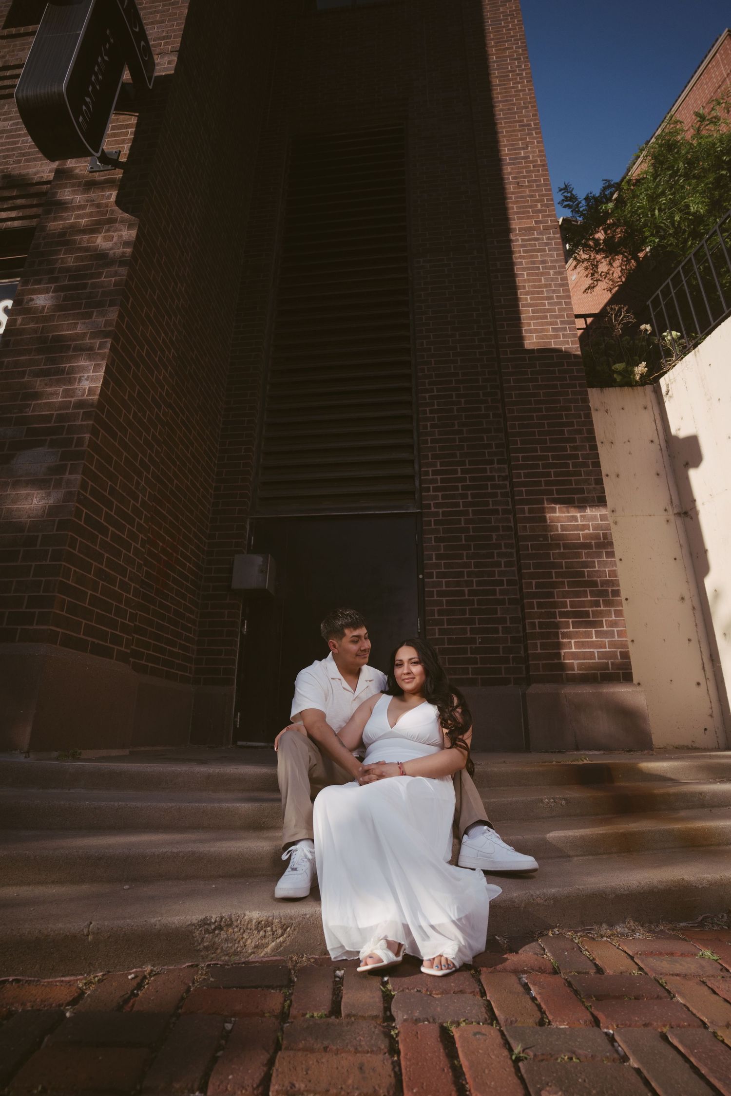 Couple in white outfits sitting on brick steps outside a building, woman in flowing dress leaning back against man.