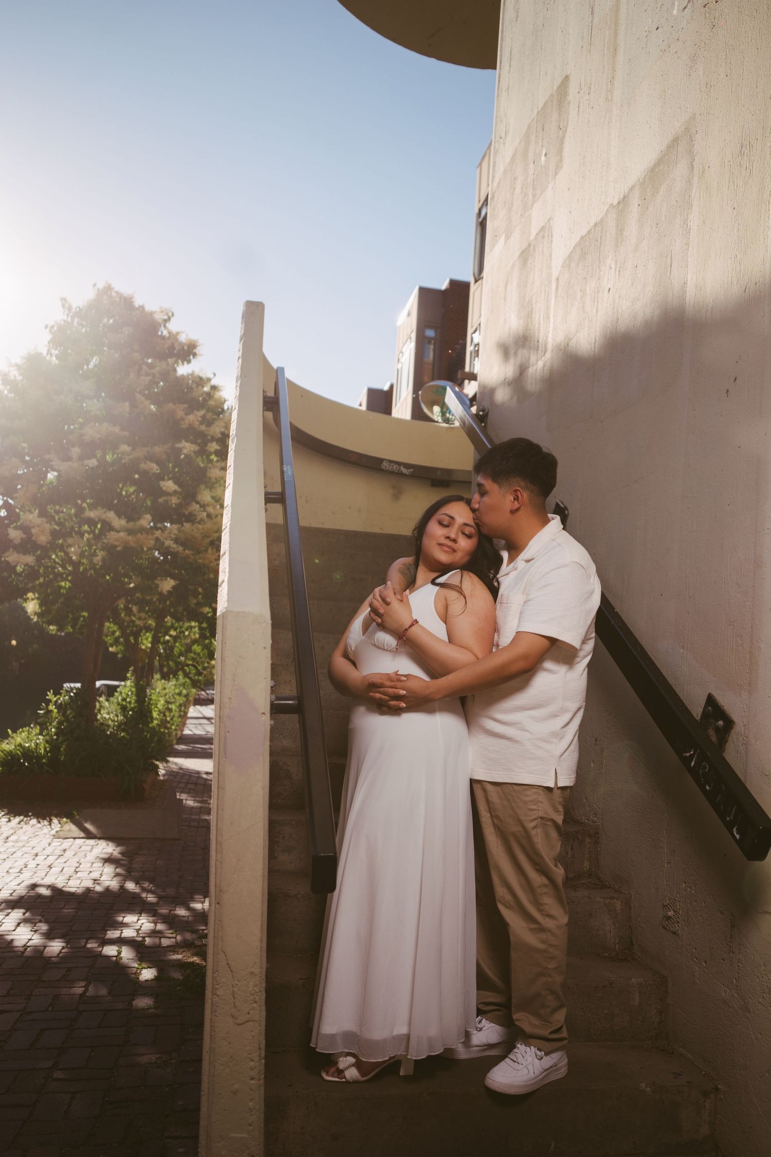 Couple embracing on urban stairs, woman in white dress, man in white shirt, backlit by golden sunlight.