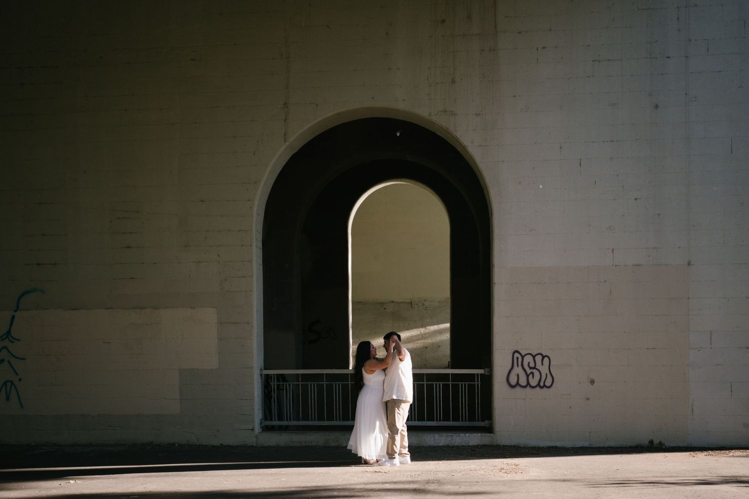 Couple embracing under a grand arched doorway of a white brick building with graffiti in soft natural light.
