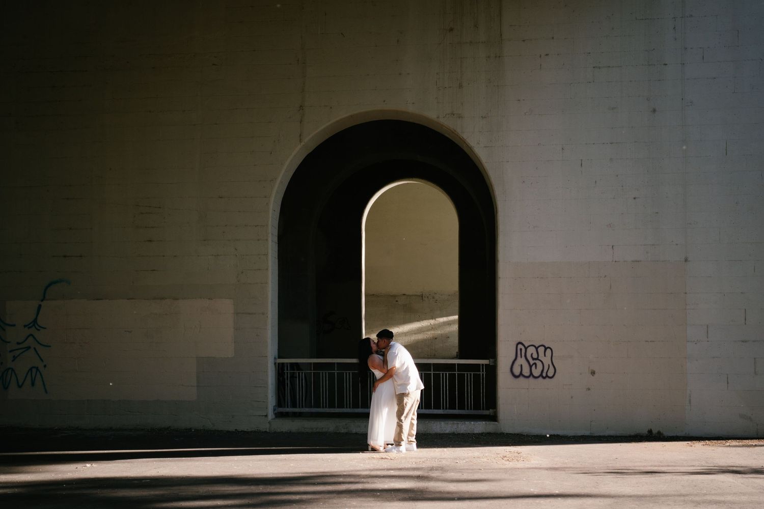 Couple shares romantic kiss under dramatic stone archway with graffiti in urban setting.