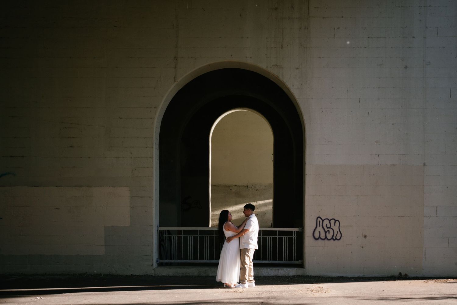 Couple sharing a romantic kiss under a grand arched bridge, surrounded by sunlight and urban architecture.