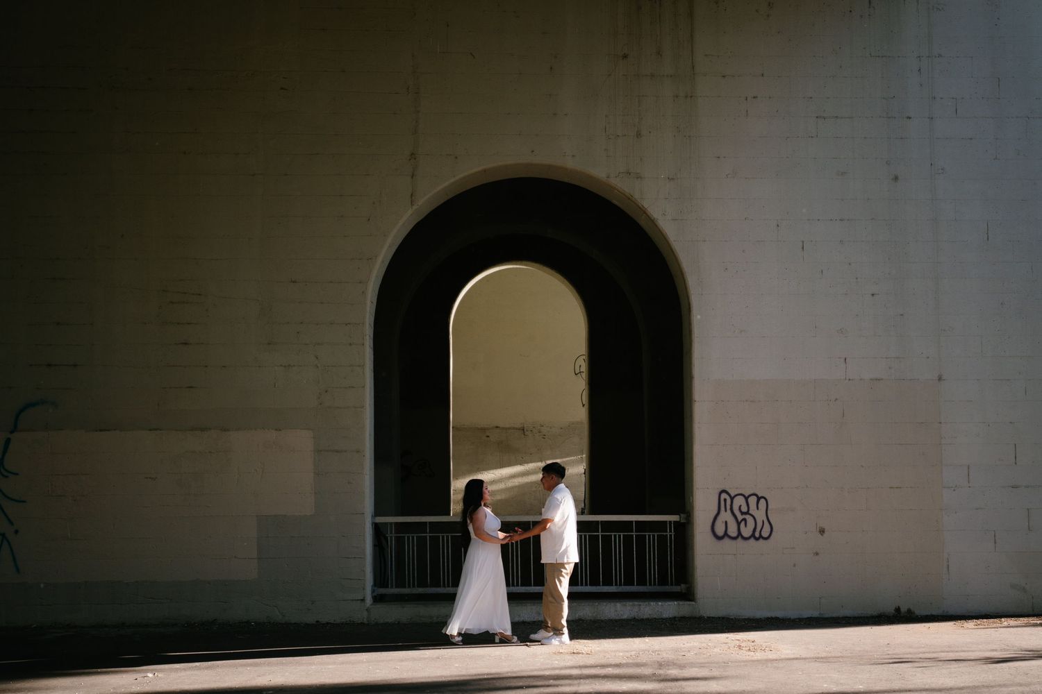 Couple holding hands under a large stone arch bridge, bathed in warm sunlight during an engagement shoot.