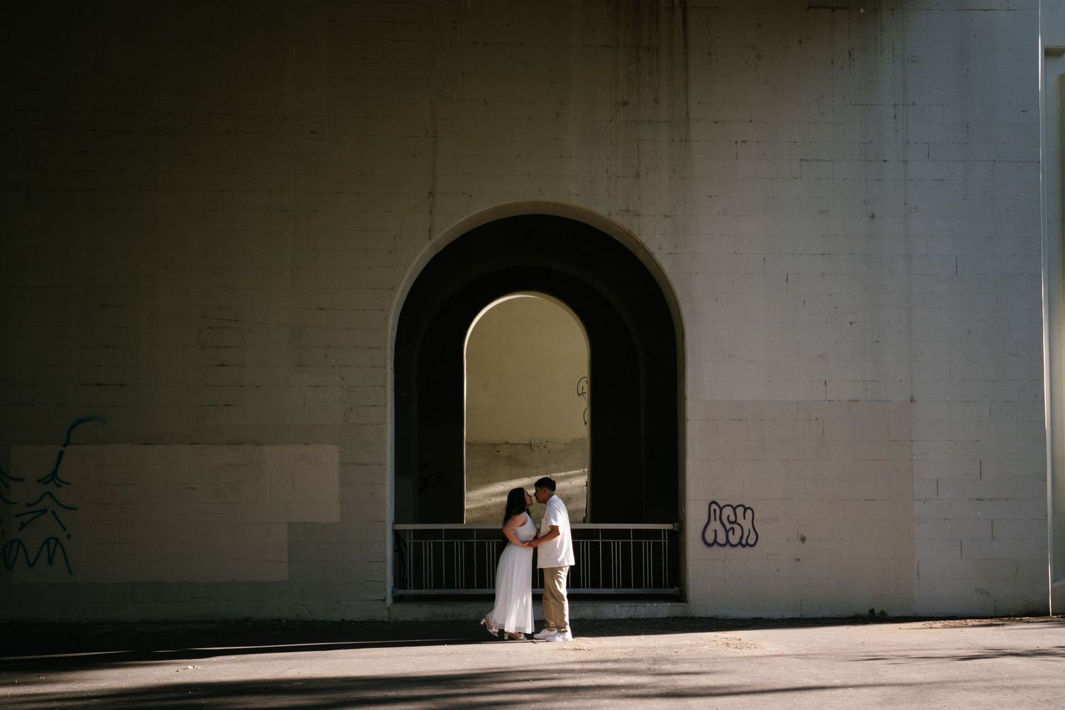 Wedding couple sharing an intimate moment framed by a dark arched doorway on a sunlit white brick wall.