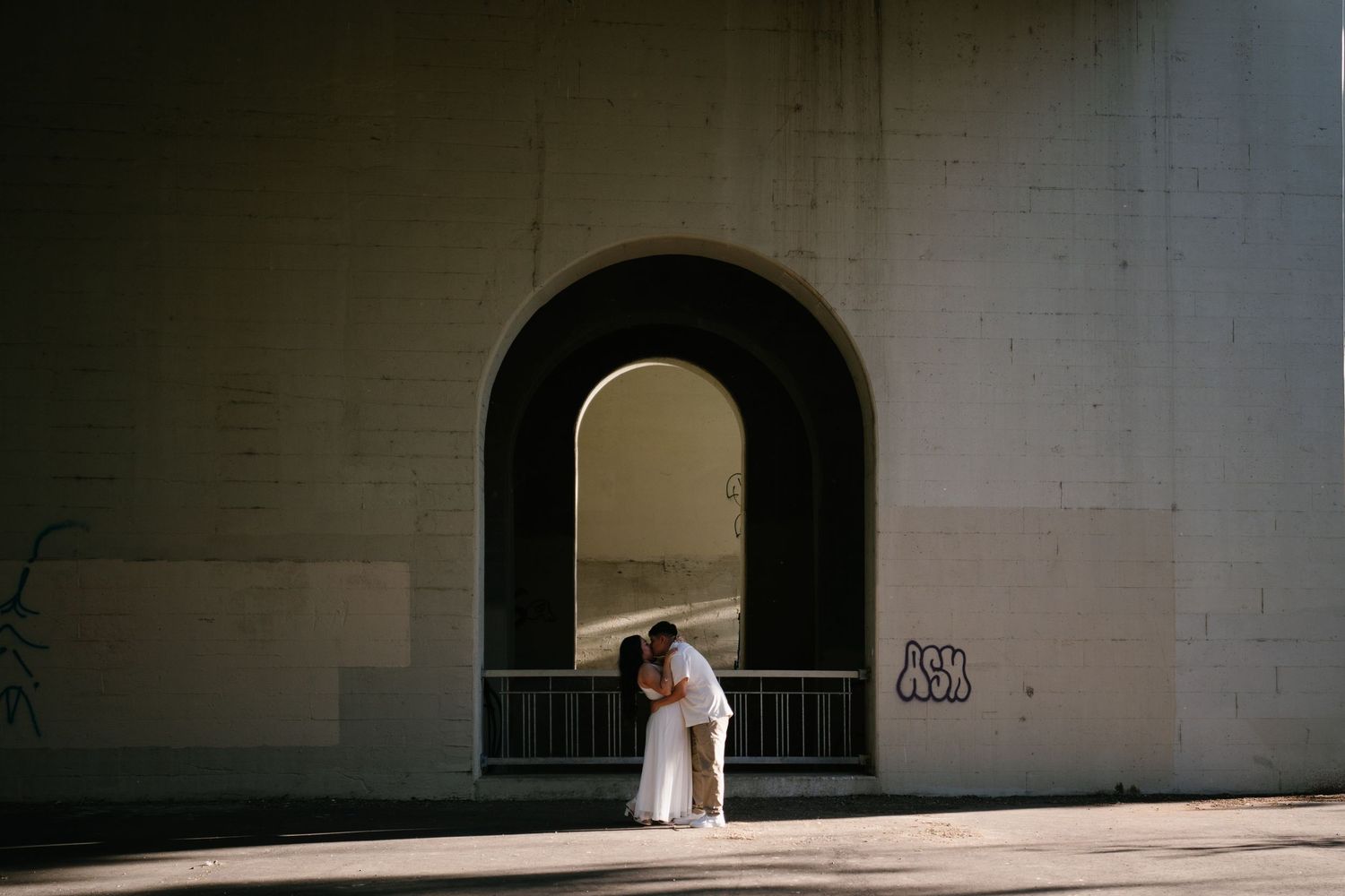 Couple sharing a kiss under a large arched bridge tunnel with graffiti on sunlit walls.