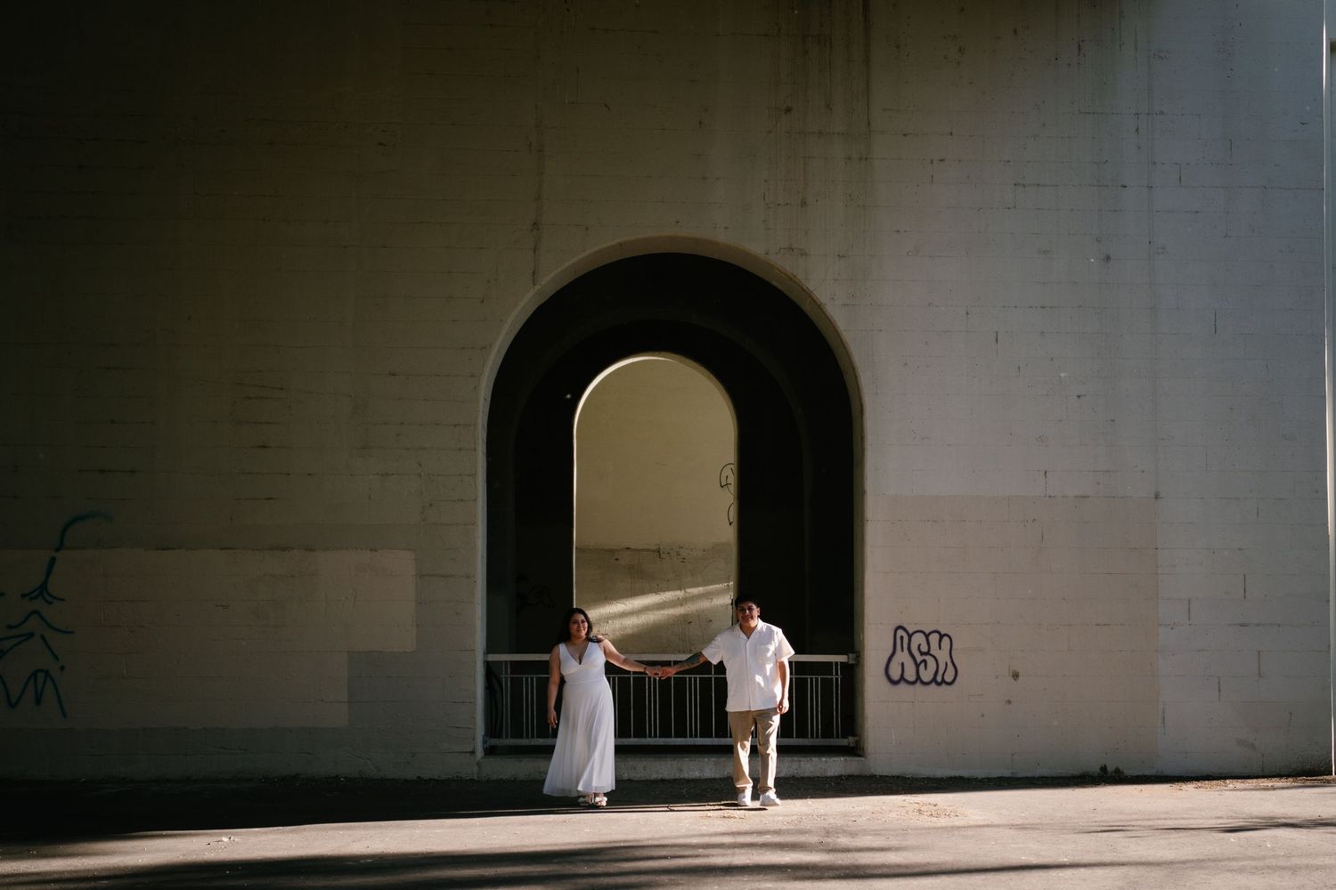 Couple in white attire standing under a large stone arch bridge with graffiti, bathed in natural sunlight.