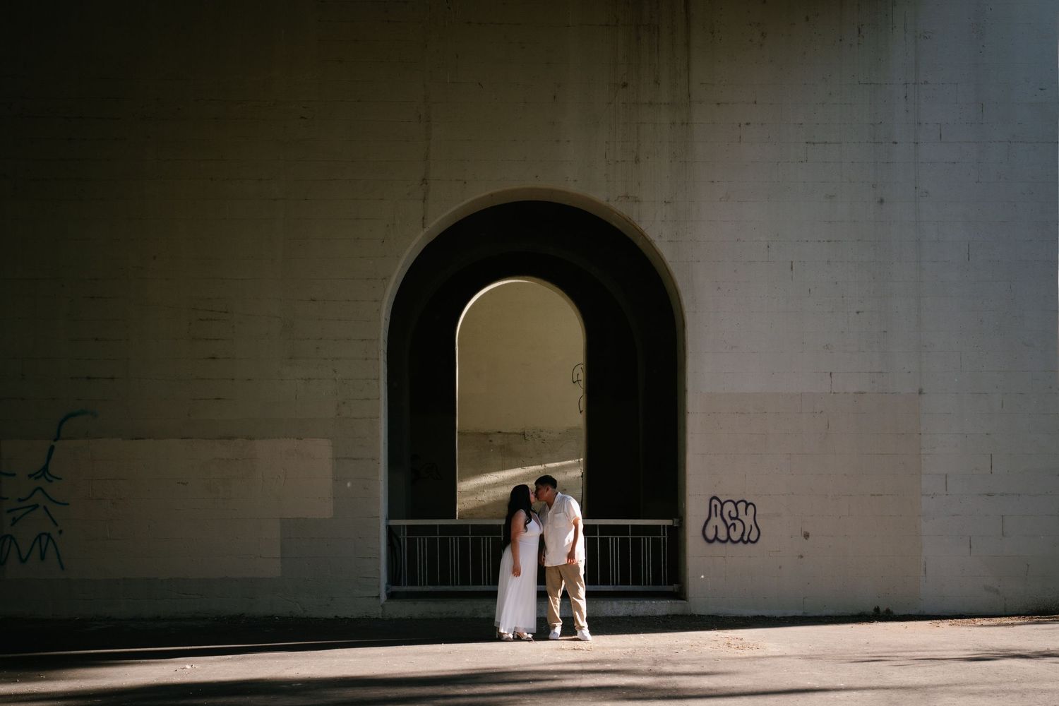 Couple standing together under a large arched bridge with graffiti in soft natural light.