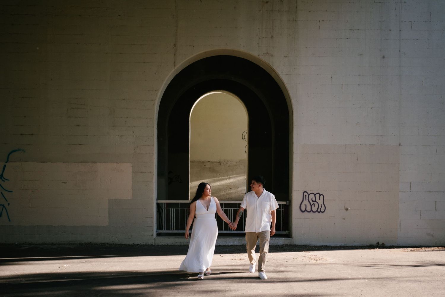 Couple holding hands walking through a large arched tunnel with sunlight streaming through.