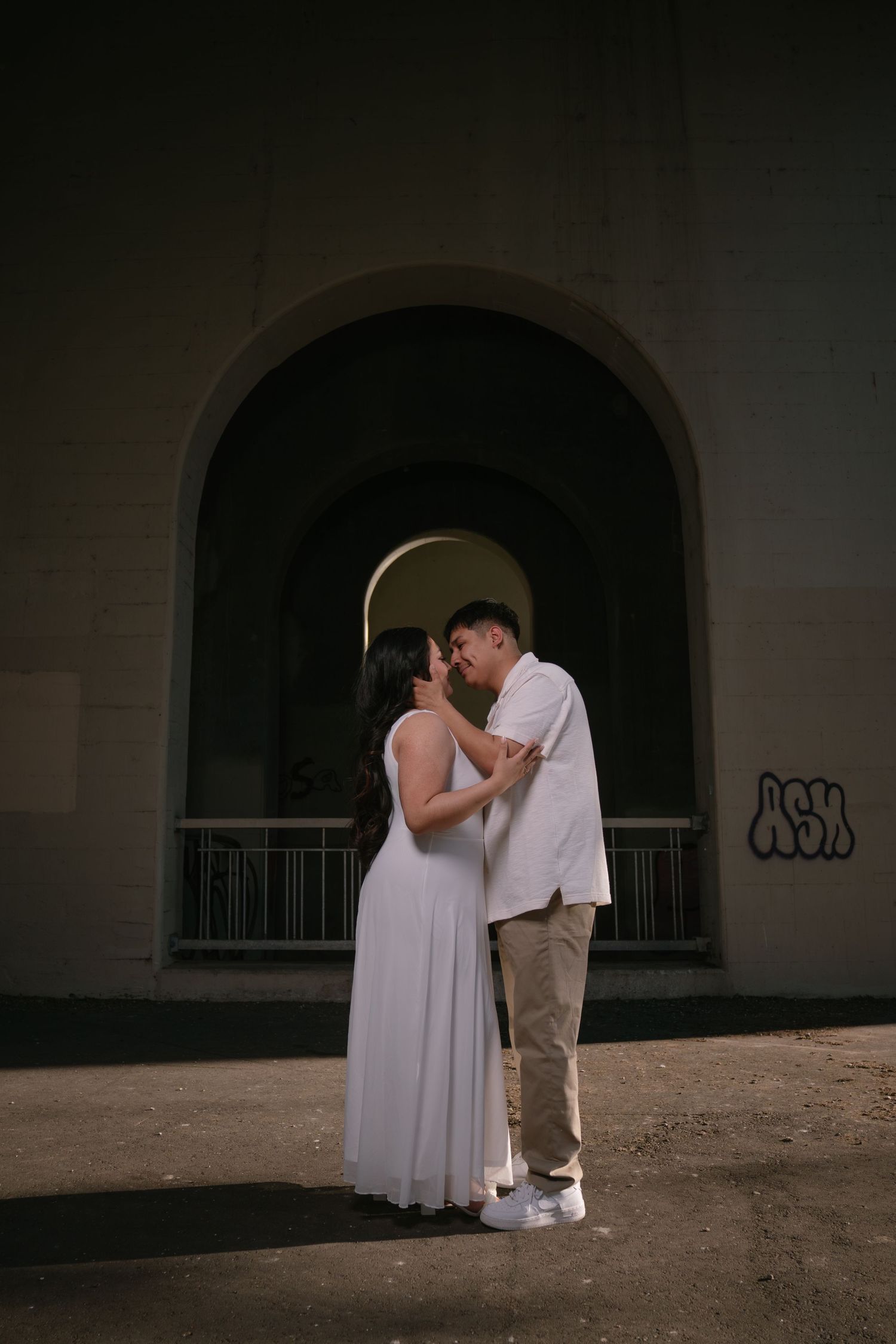 Couple sharing a tender kiss in front of an arched wall with graffiti in dramatic lighting.