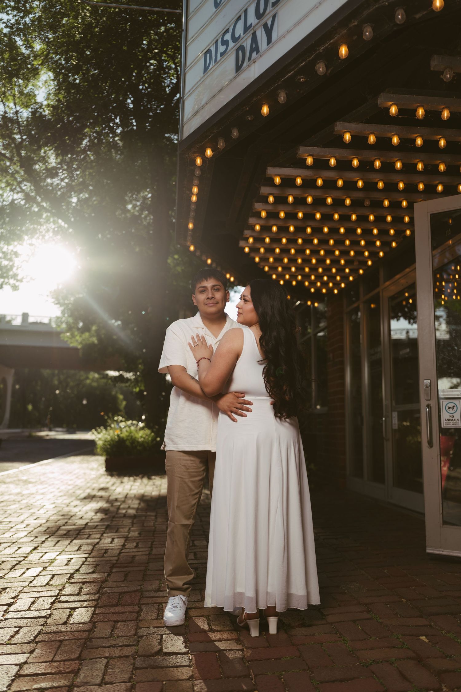 Couple embracing under a vintage marquee with glowing bulb lights outside a theater at sunset.