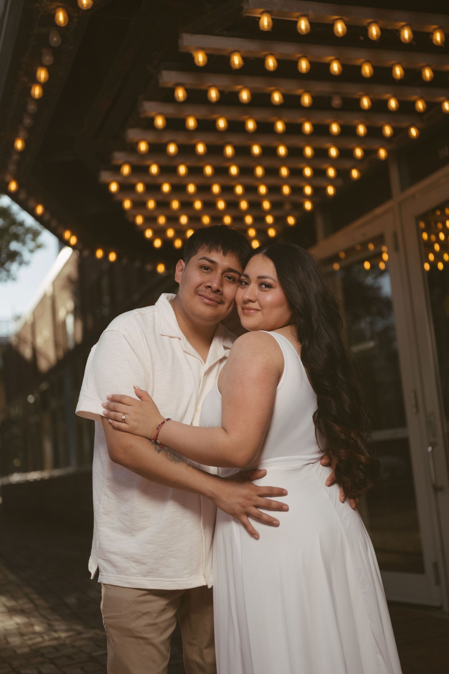 Couple hugging closely under warm string lights outside a venue entrance at dusk.