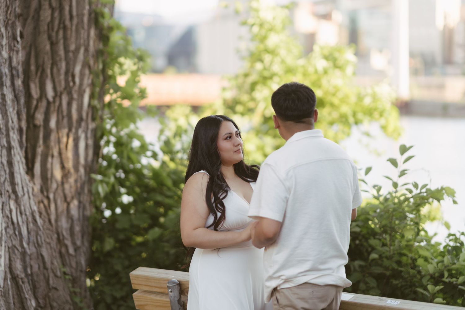 Woman in white dress smiling at her partner near a tree with a river in the background.