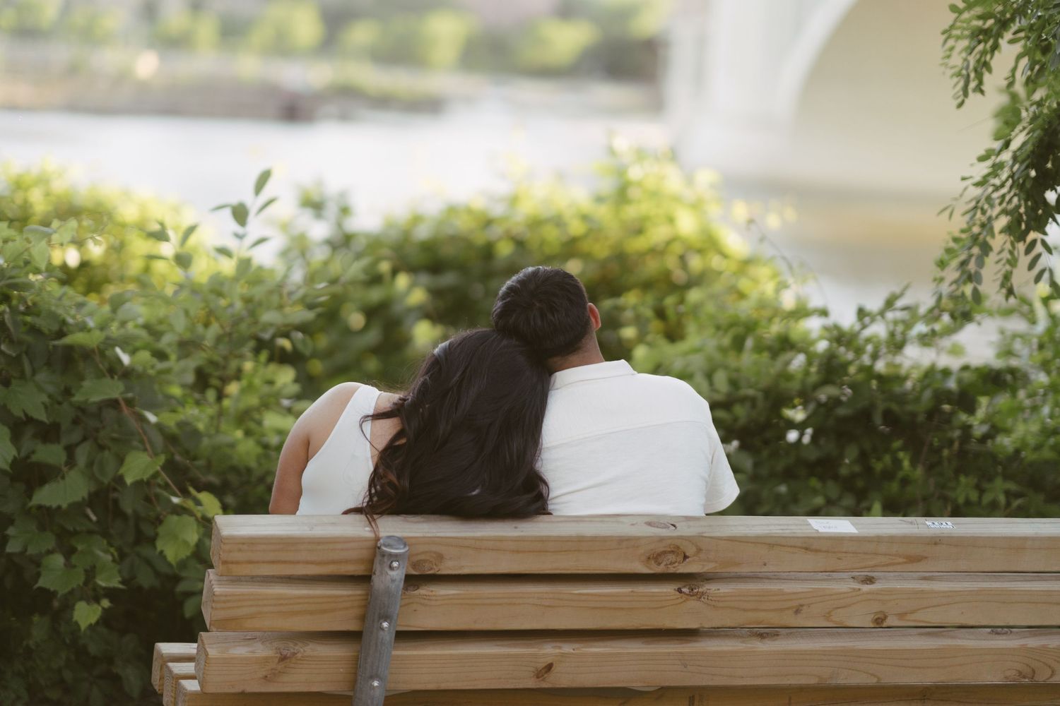 Couple sitting close together on a wooden bench surrounded by lush green foliage near water.