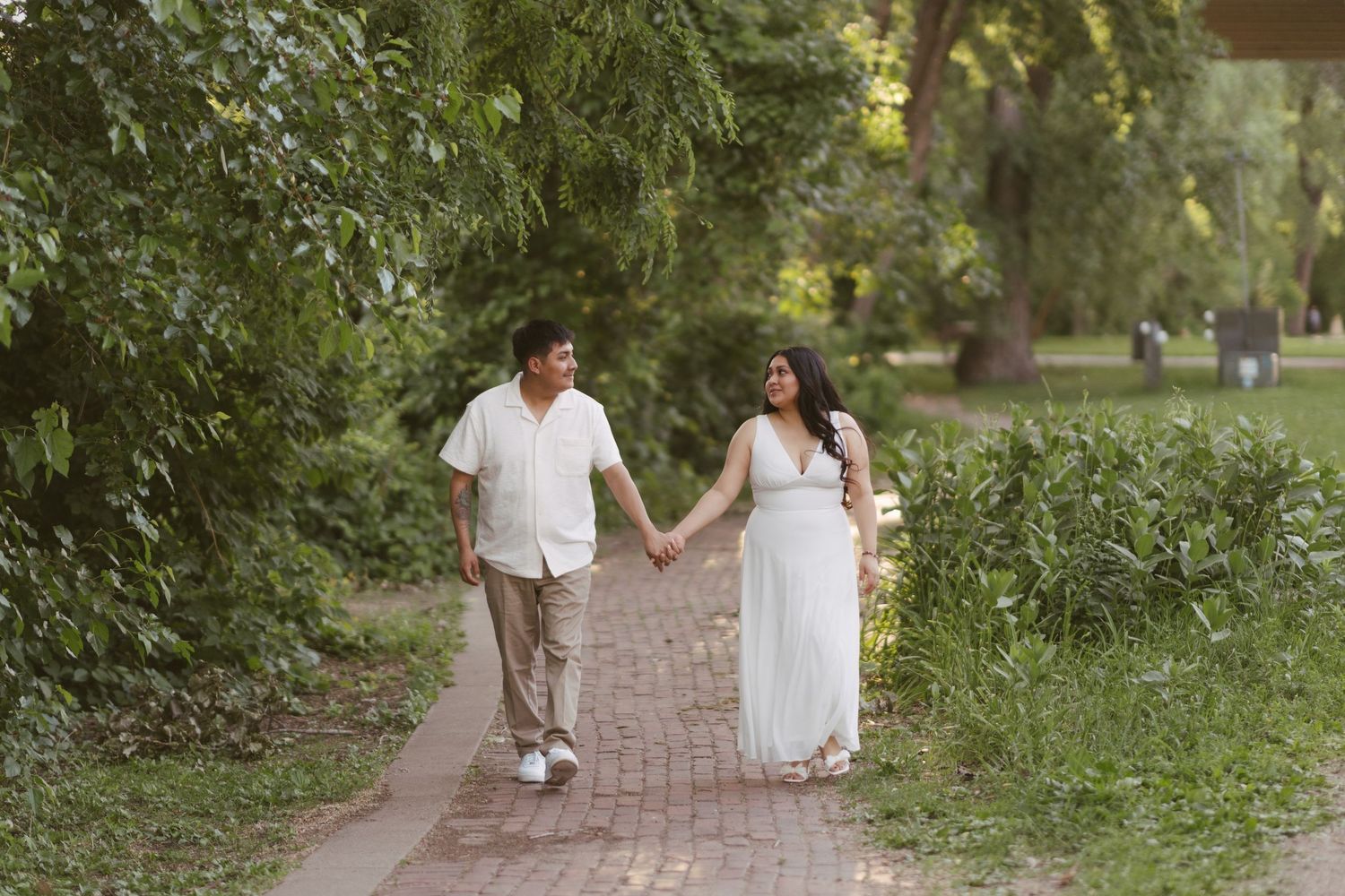 Couple holding hands walking on brick path through lush green park, woman in white maternity dress.