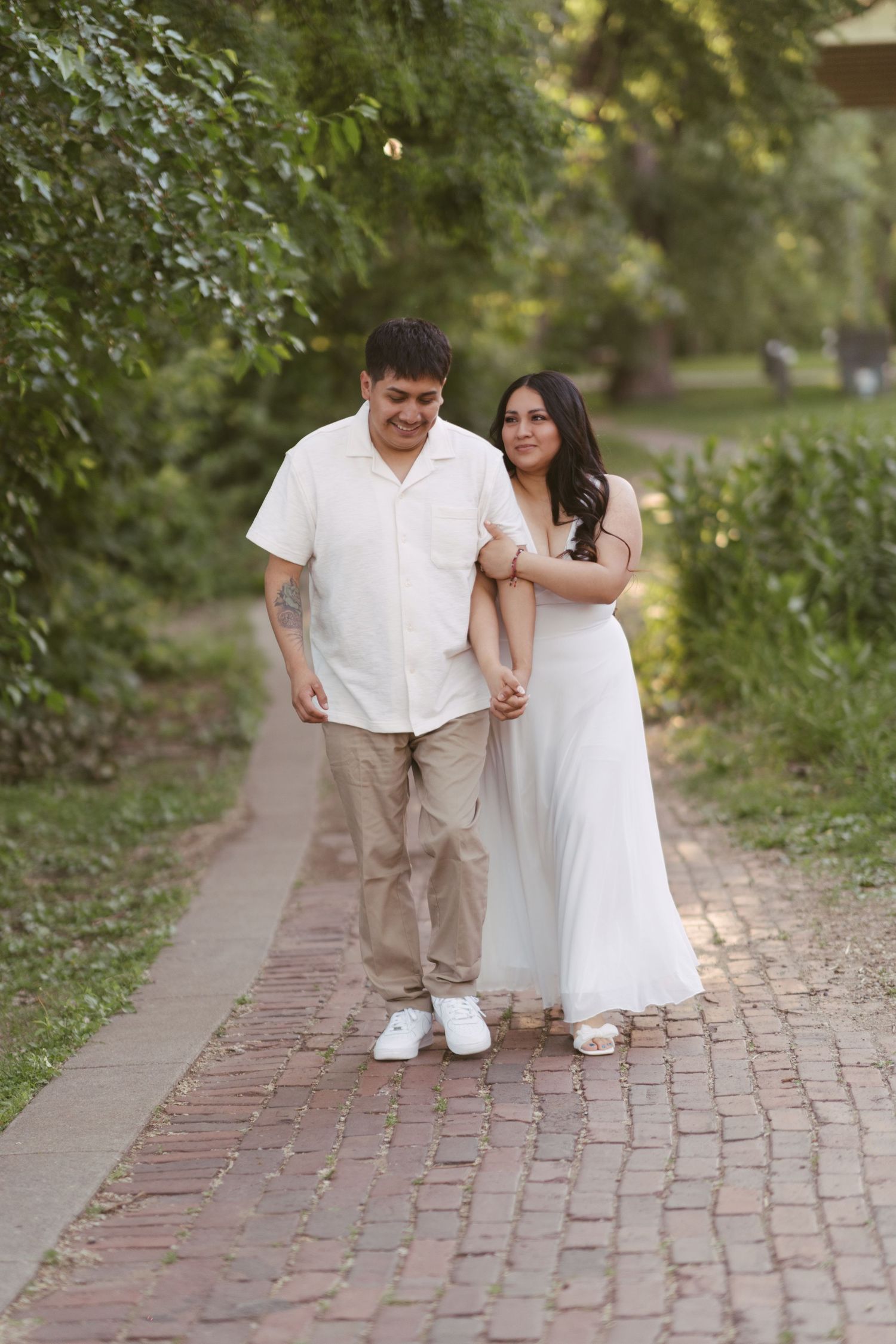 Smiling couple walking hand-in-hand along a brick path surrounded by lush green trees.