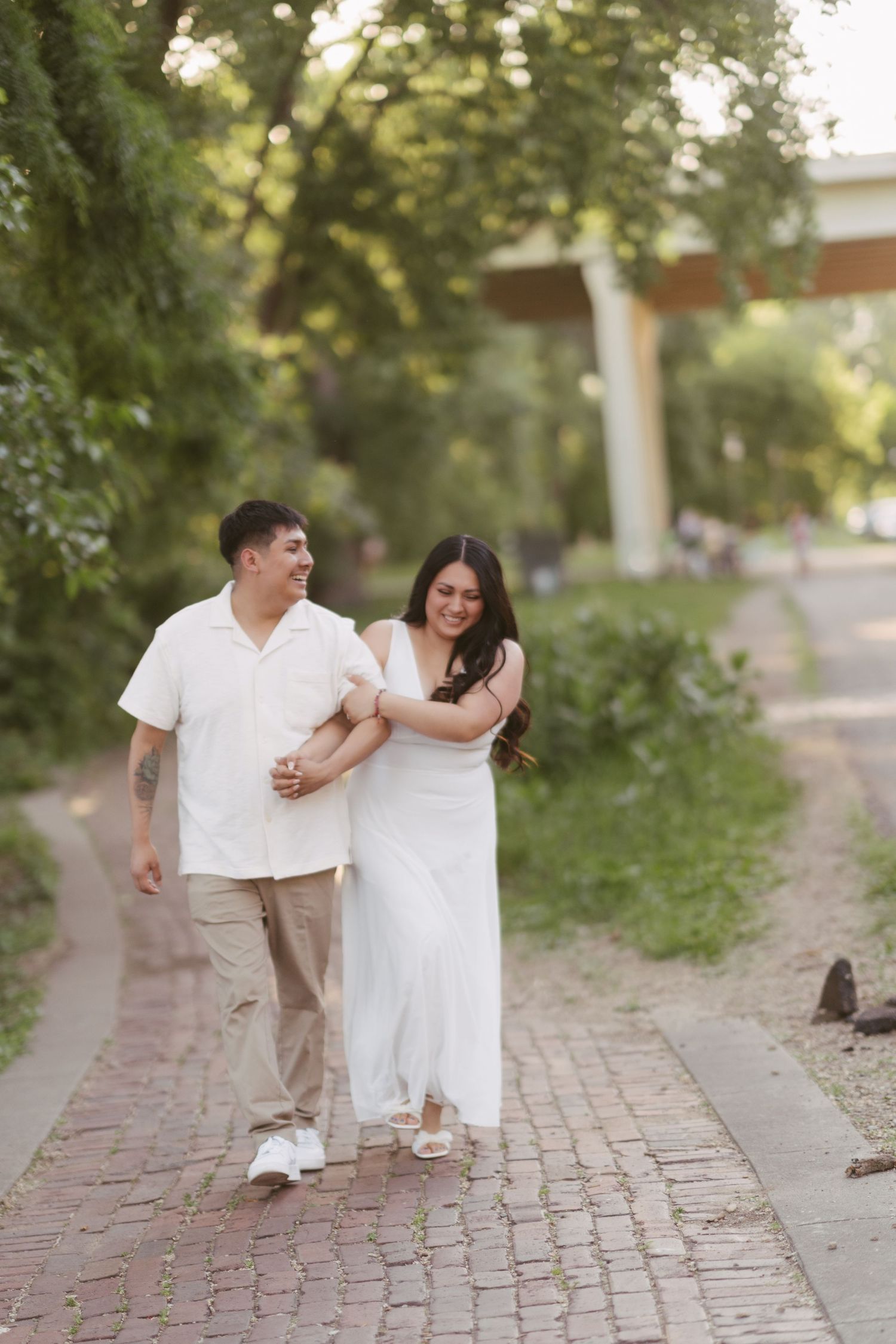 Happy couple walks arm-in-arm along brick path surrounded by lush green trees during outdoor engagement photo session.