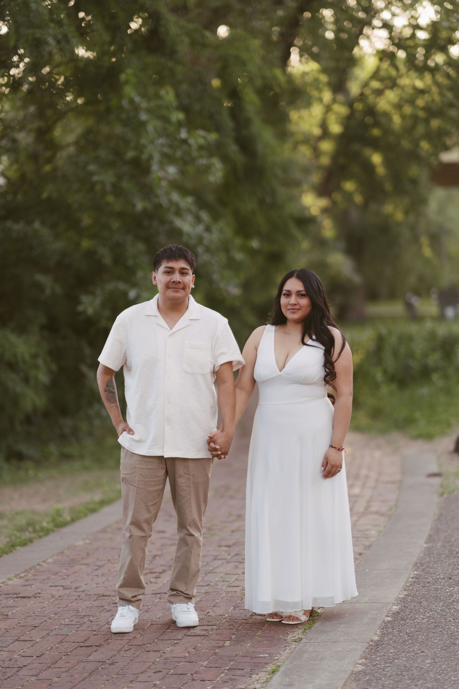 Couple holding hands and posing on a brick pathway with green trees in the background.