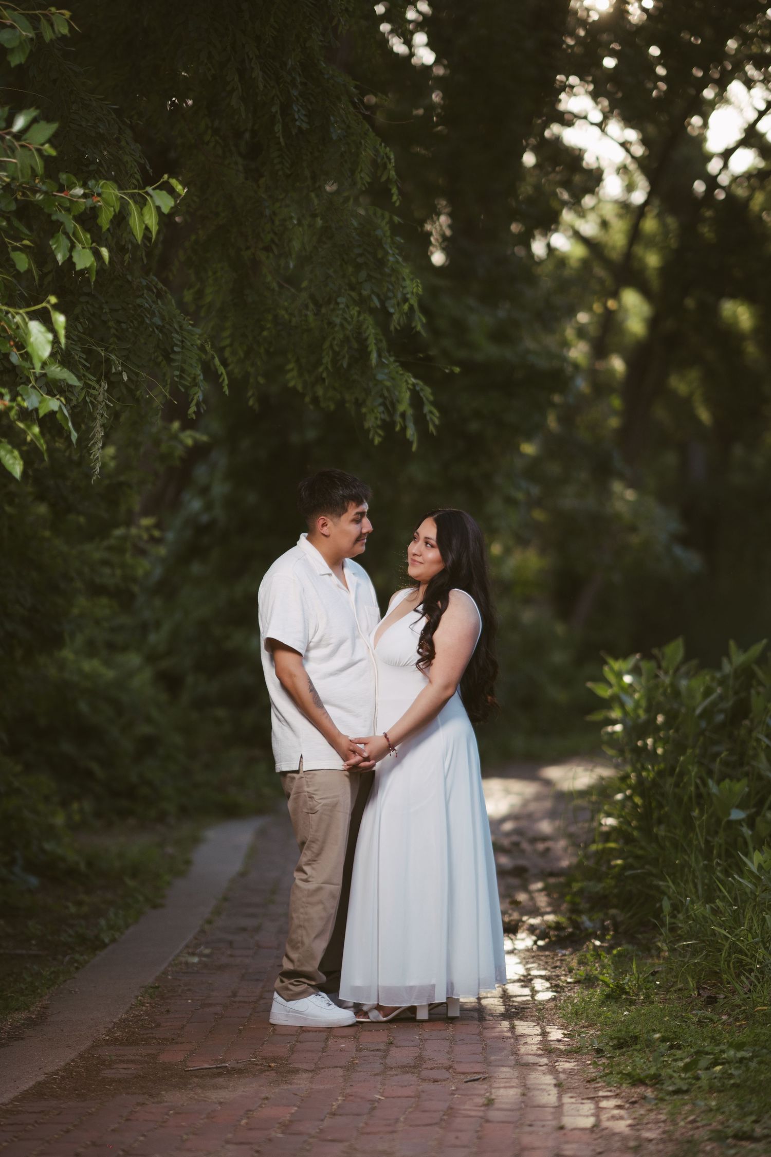 Couple holding hands on a brick garden path surrounded by lush green trees during golden hour.