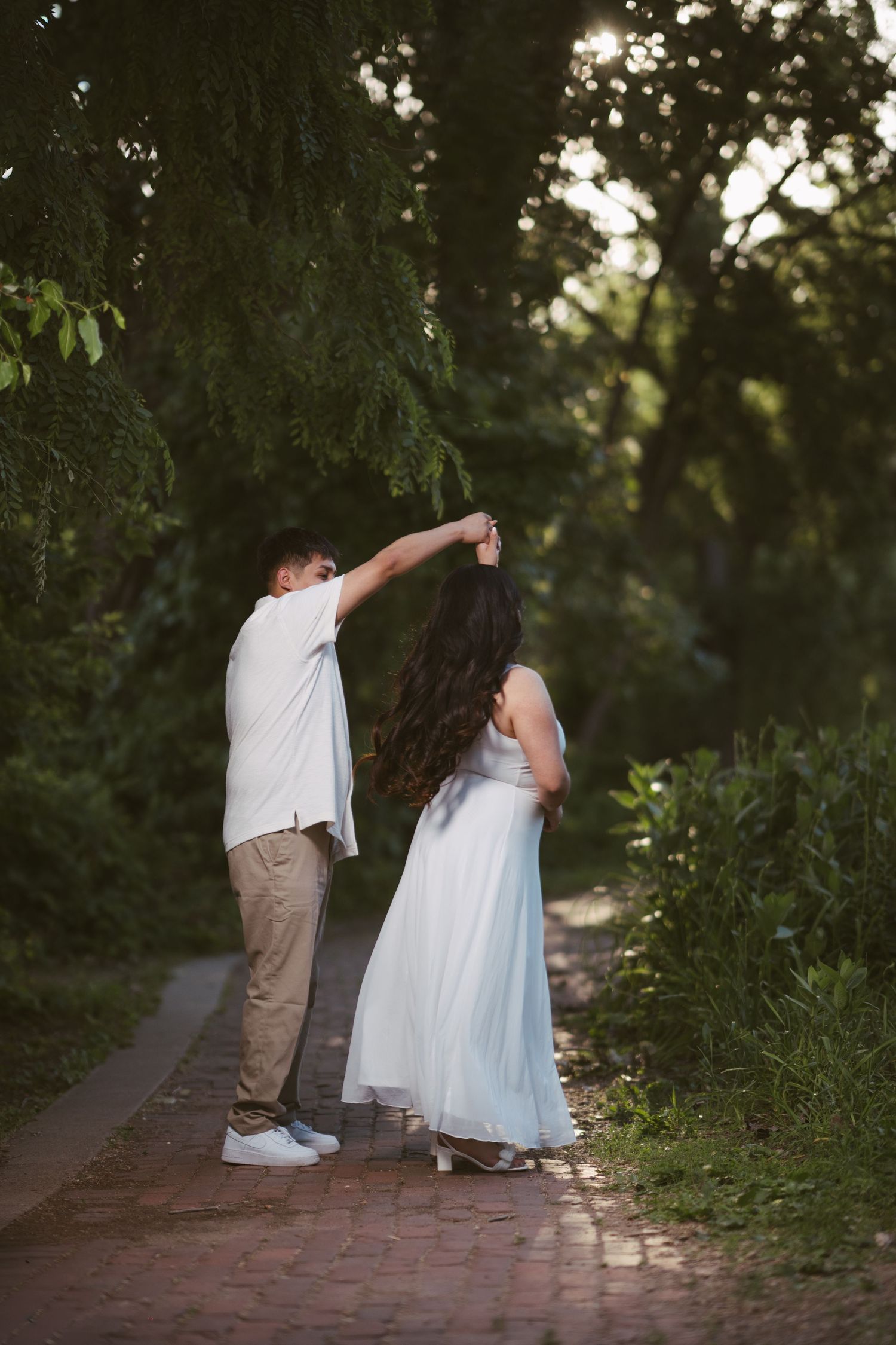 Couple dancing on a brick path in a lush green park, woman in a flowing white dress being twirled by her partner.