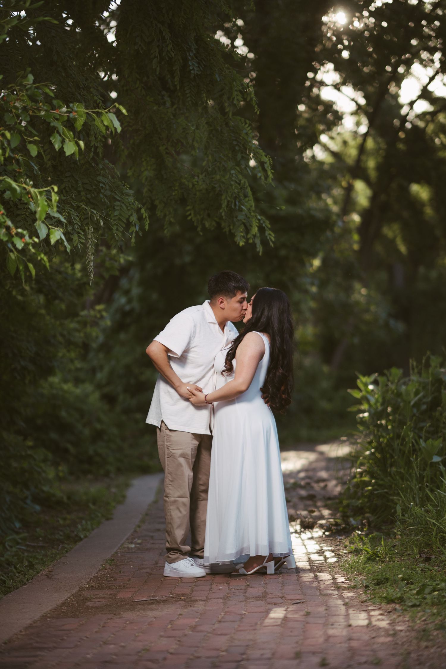 Couple kissing on a sunlit brick path surrounded by lush green trees in a park.