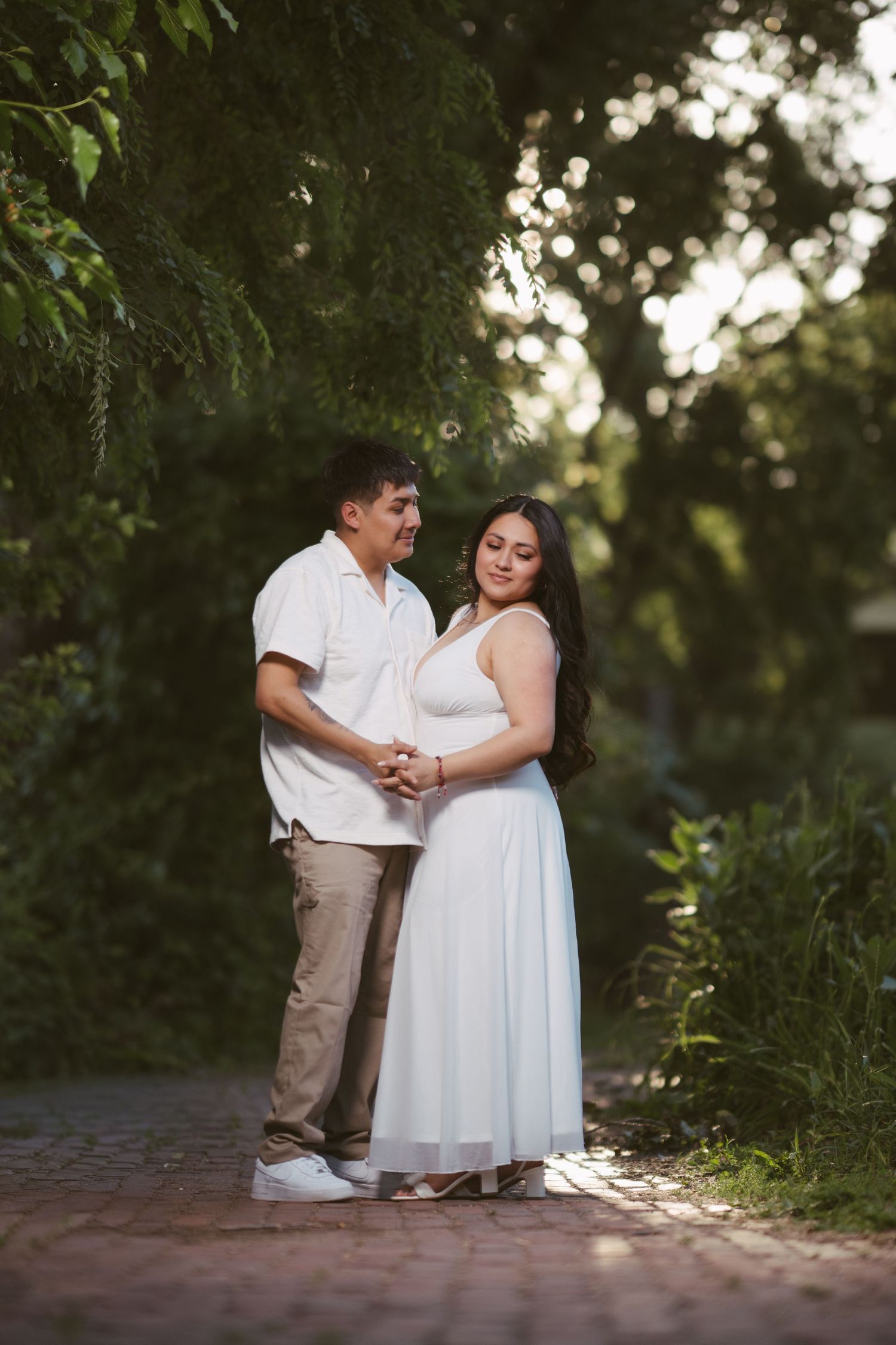 Pregnant couple holding hands on a brick path surrounded by green trees in soft golden light.