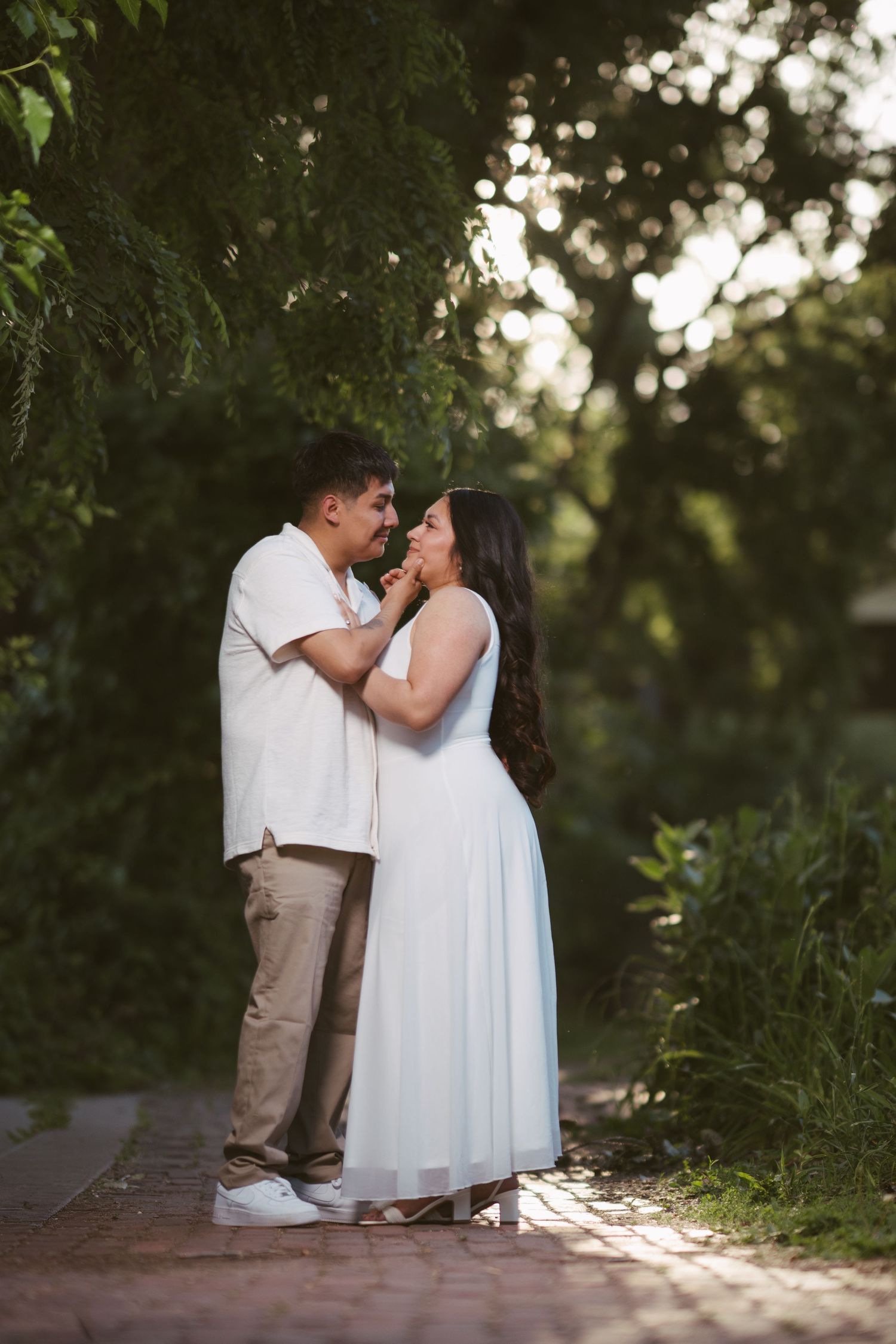 Couple sharing an intimate moment on a sunlit brick path surrounded by lush green foliage.