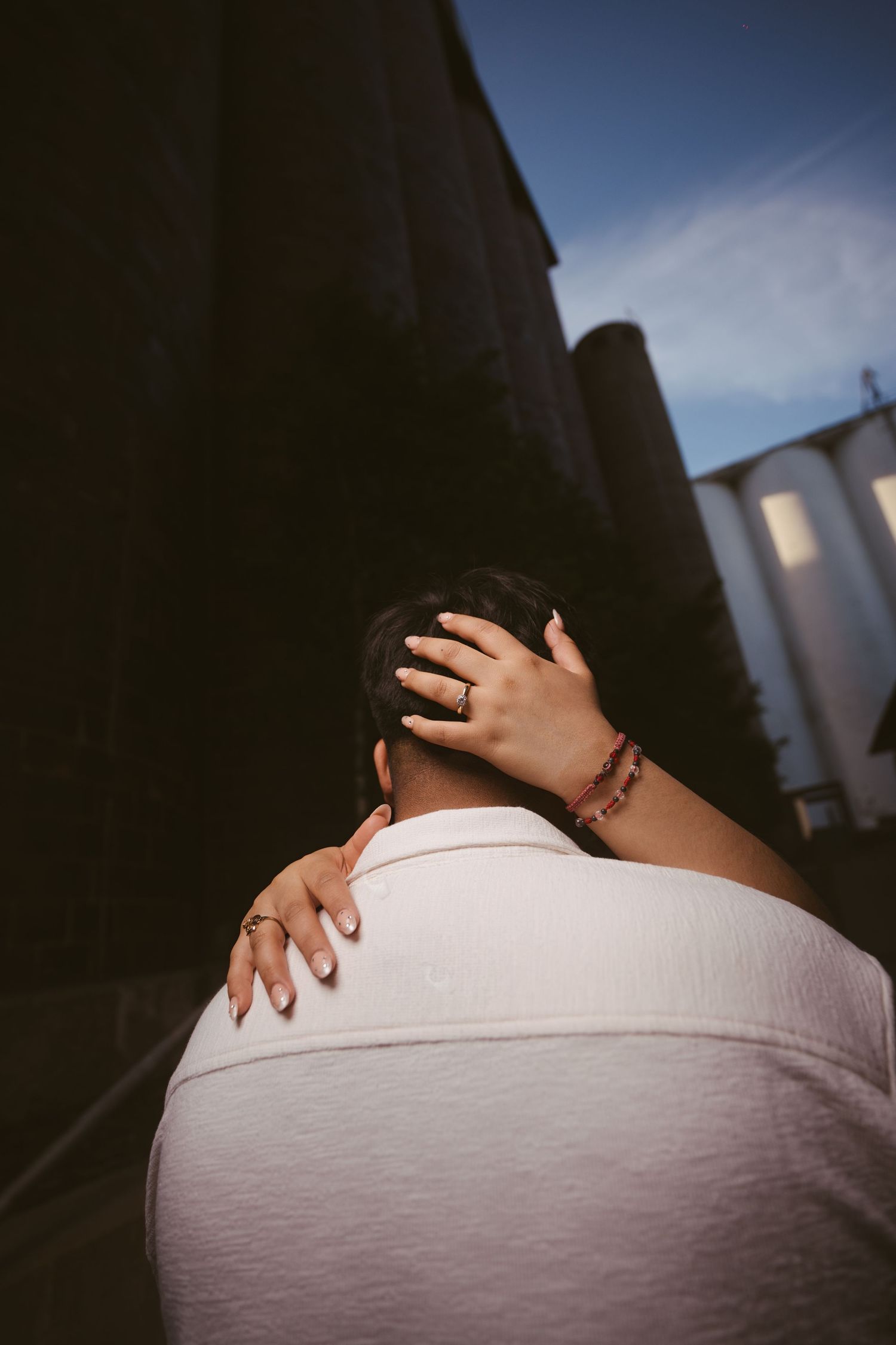 A woman rests her hand on a man's shoulder during a romantic embrace, wearing a ring and bracelet outdoors.