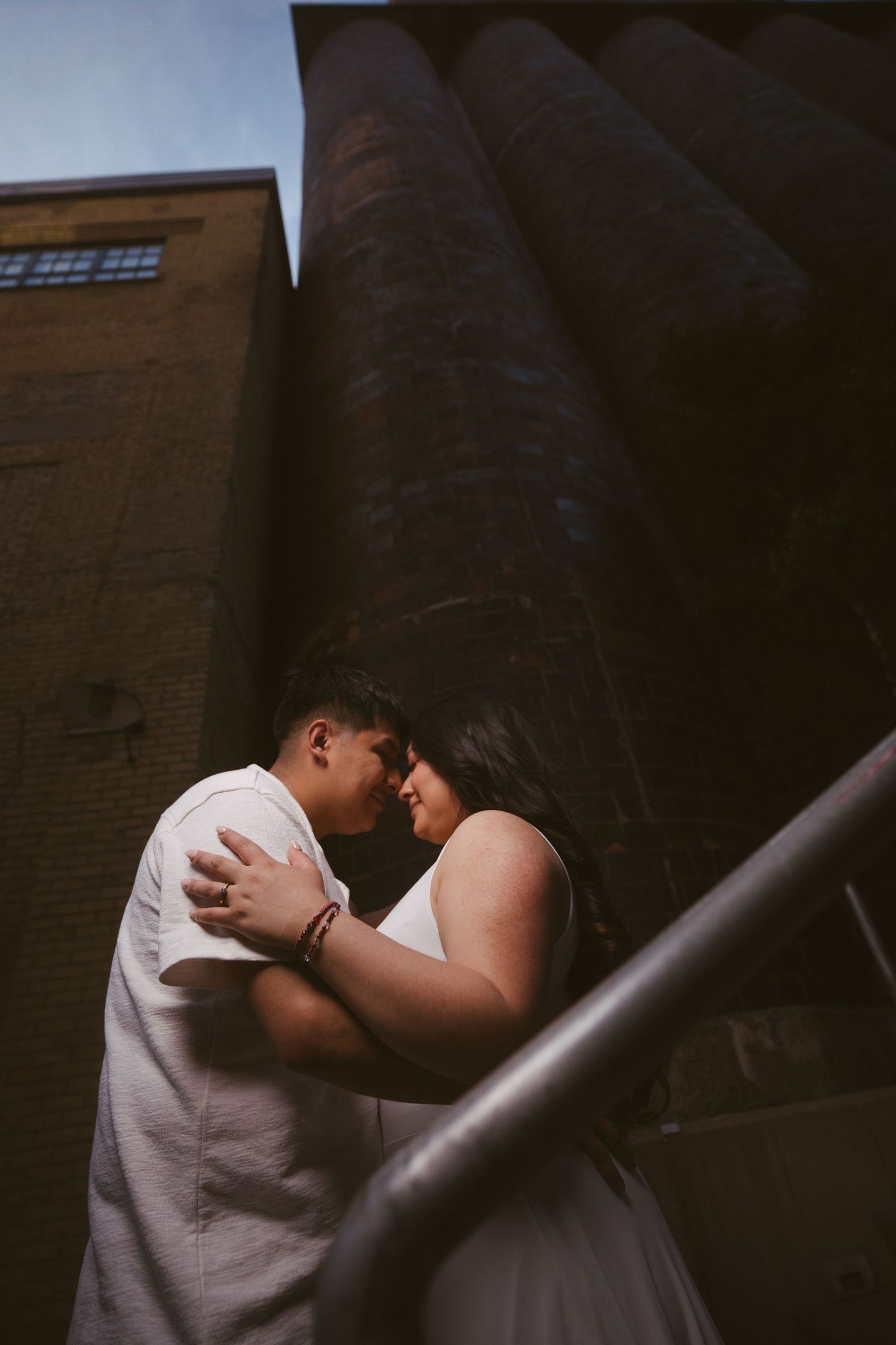 Intimate couple nearly kissing on staircase beside towering brick industrial building at dusk.