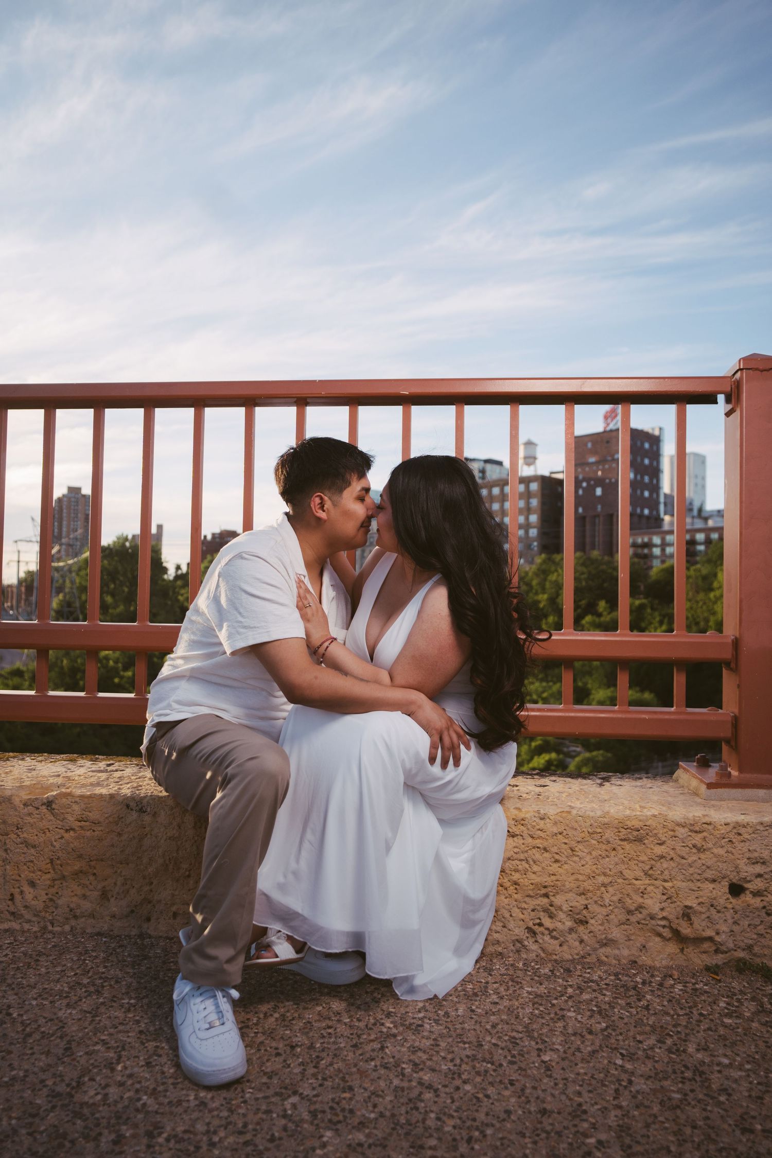 Couple sharing a kiss while sitting on a bridge with a city skyline in the background.