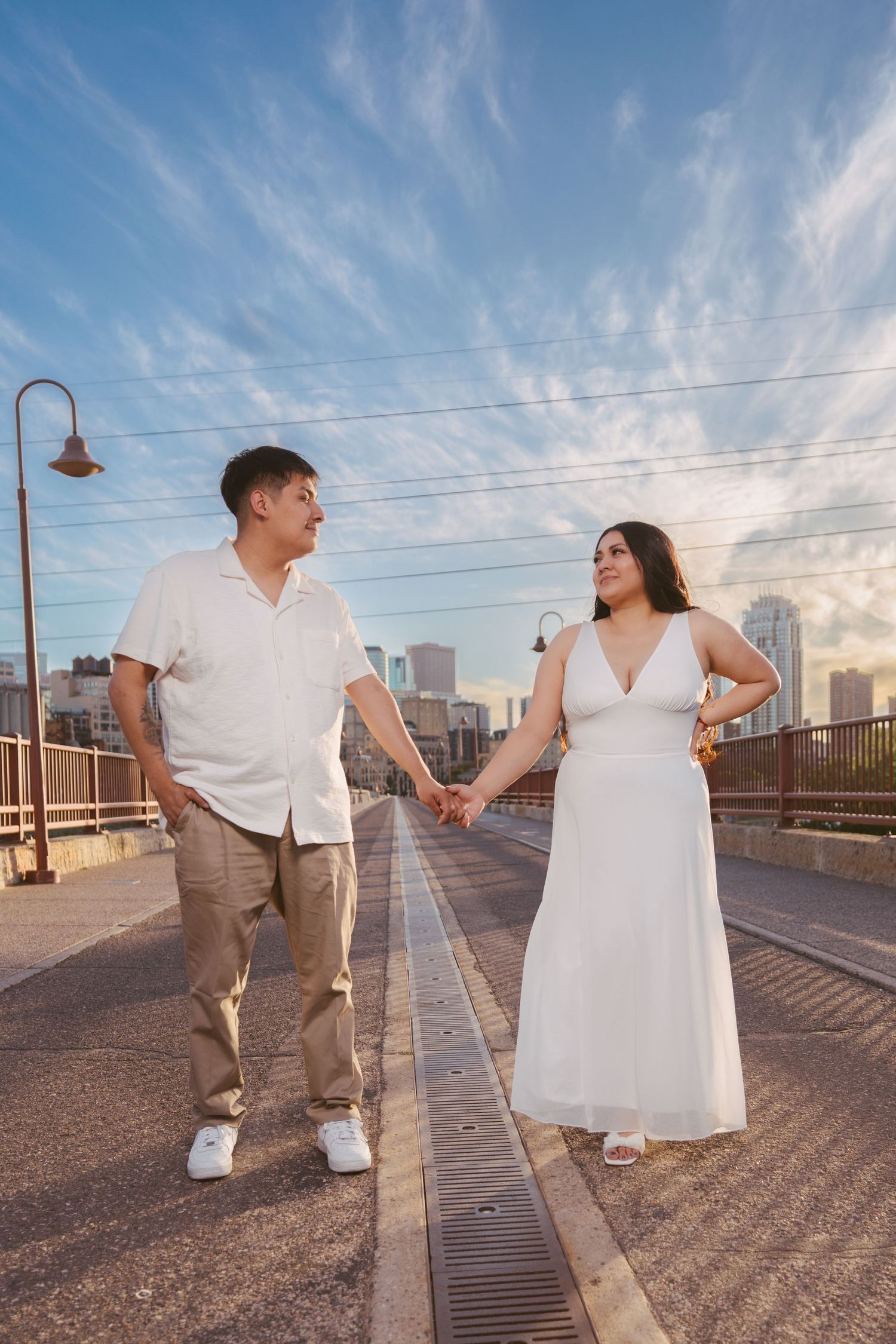 Couple holds hands walking on urban bridge with city skyline and golden hour clouds behind them.