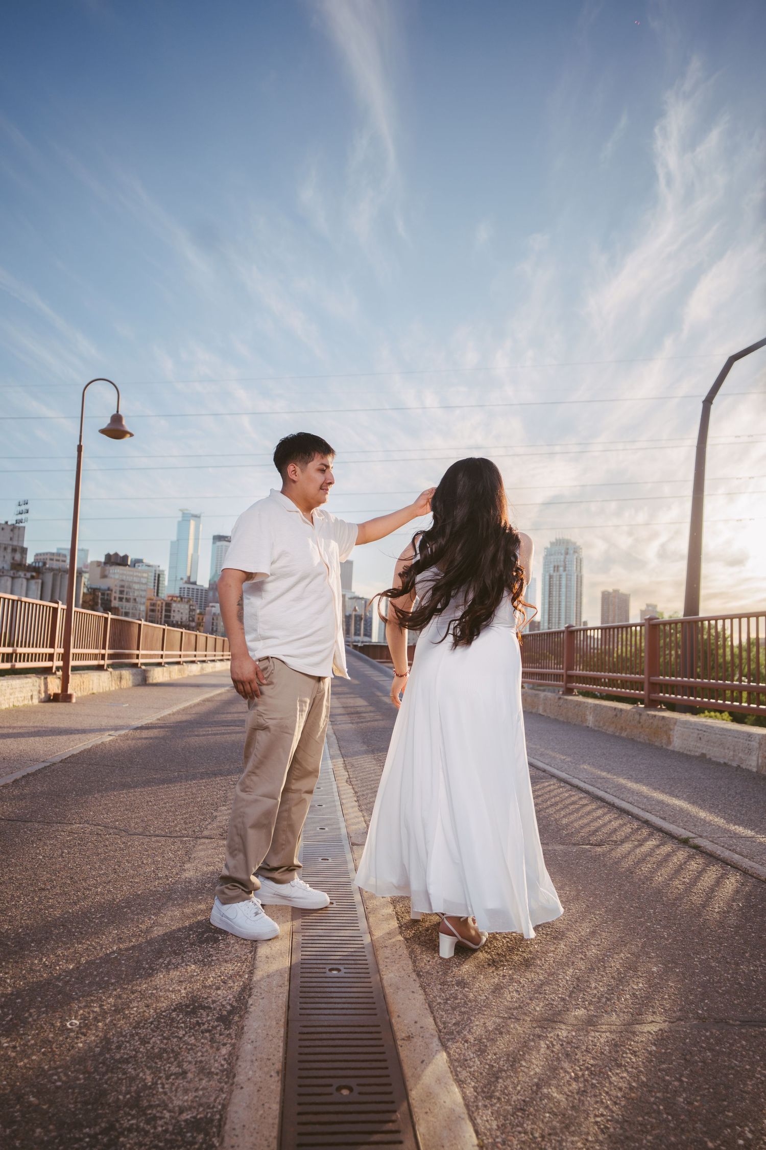 Couple dancing on a bridge at golden hour with city skyline backdrop, woman in white dress, man in white shirt.