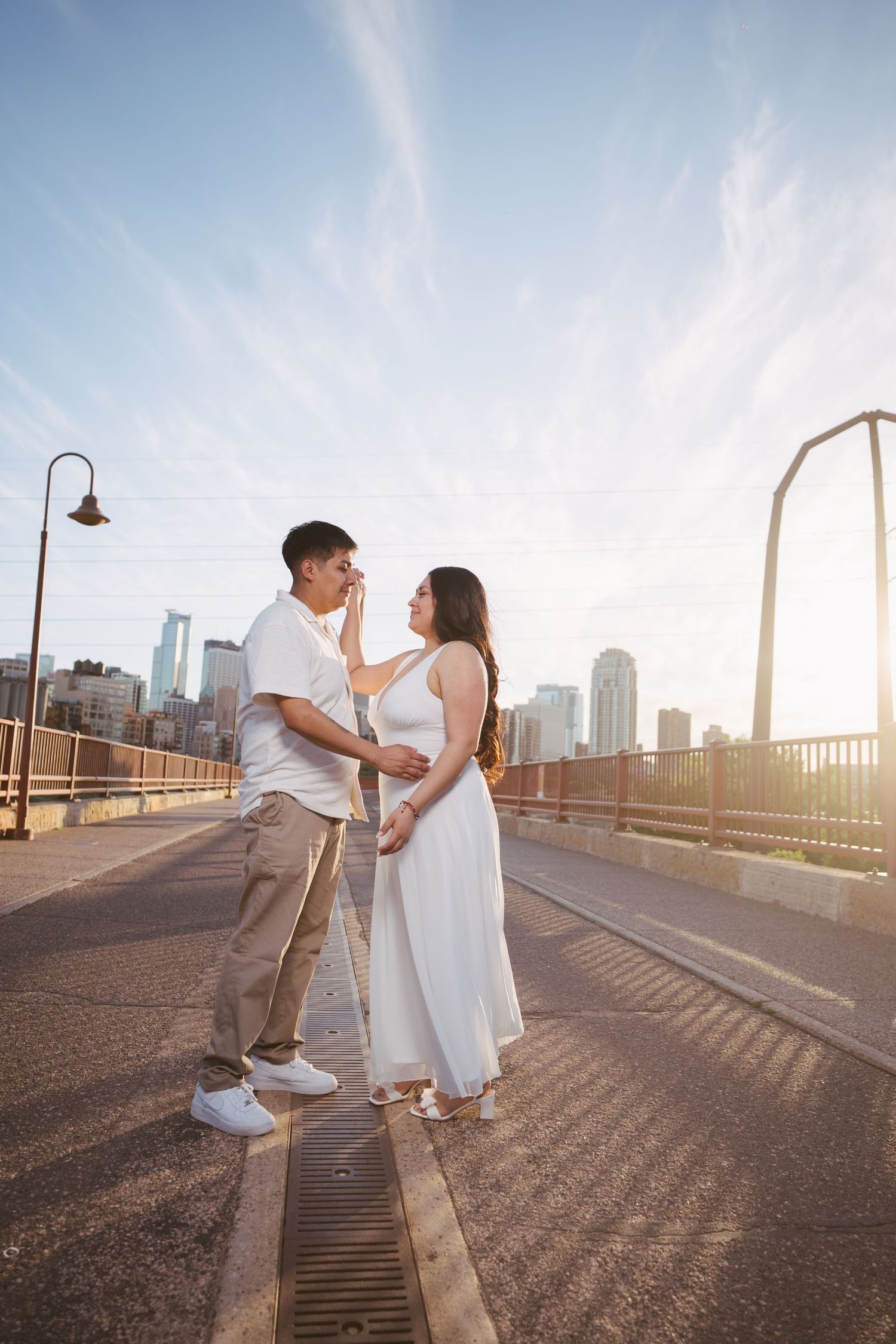 A couple shares a tender moment on a bridge at golden hour with a city skyline backdrop.