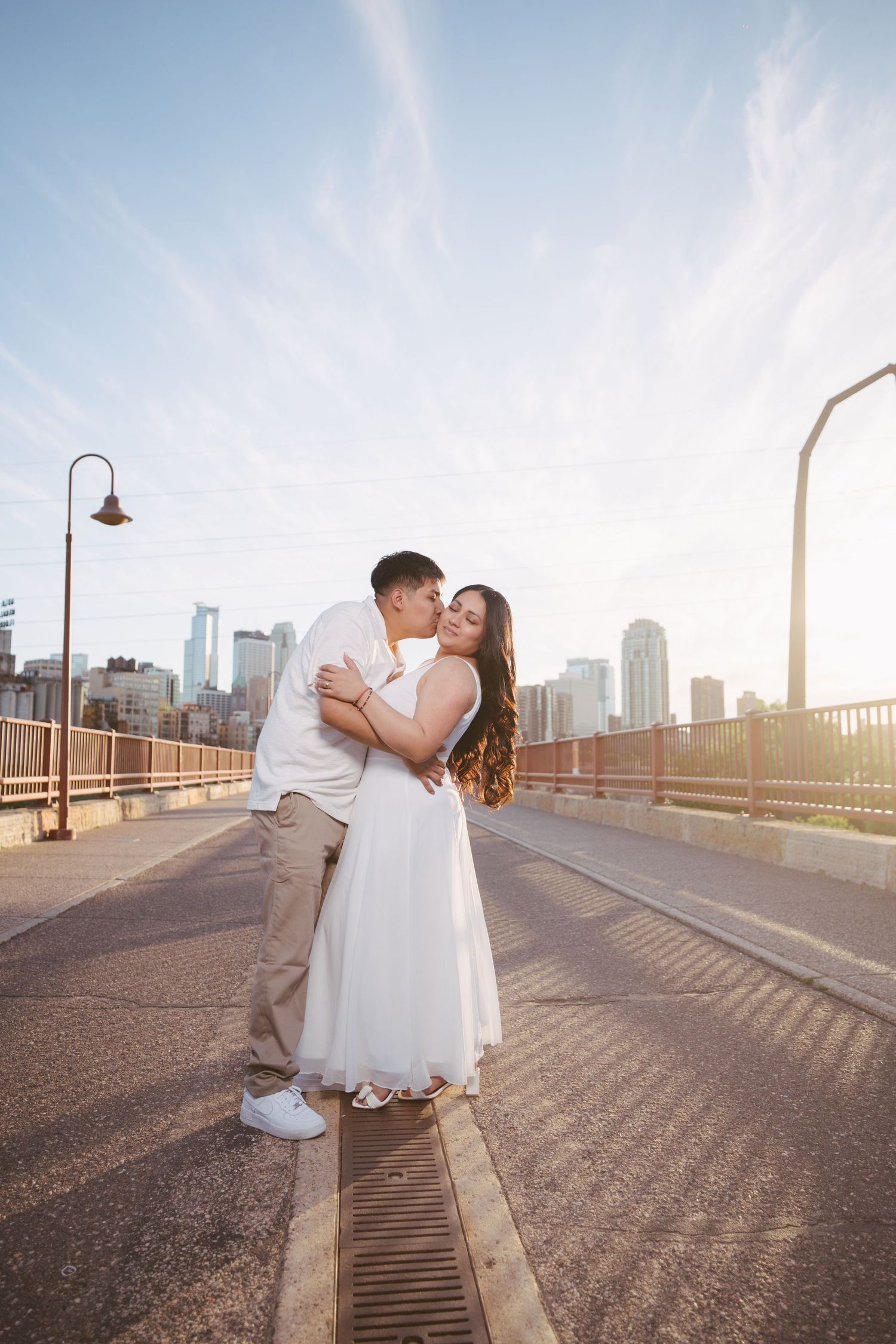 Couple embraces and kisses on sunlit bridge with stunning city skyline glowing in background.