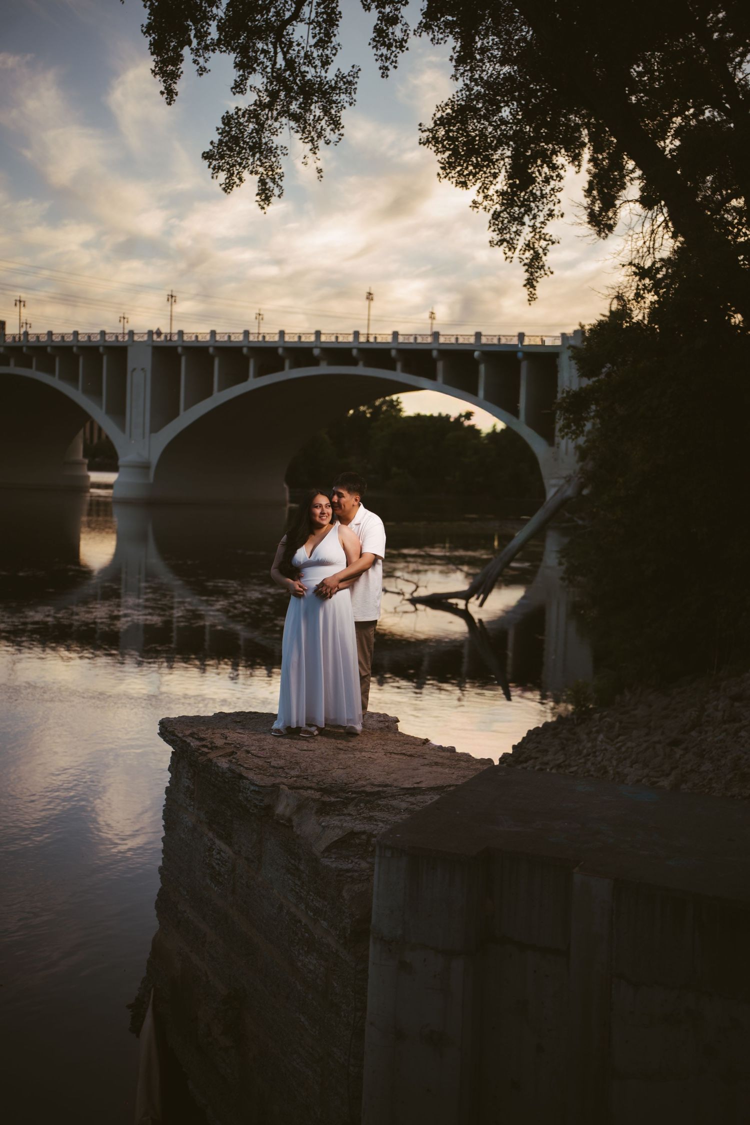 Romantic couple embracing on stone pier at dusk with arched bridge reflection on calm river waters.