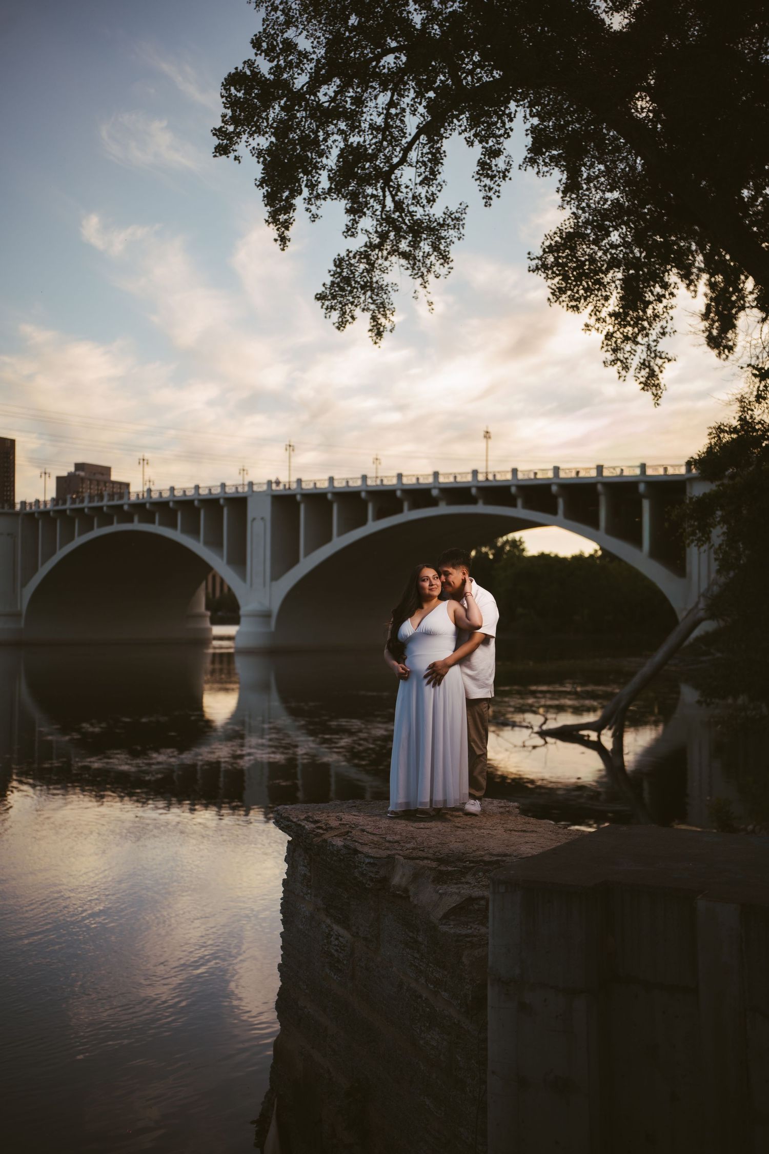 Pregnant couple embraces romantically by river at sunset with elegant arched bridge reflecting in calm water behind them.