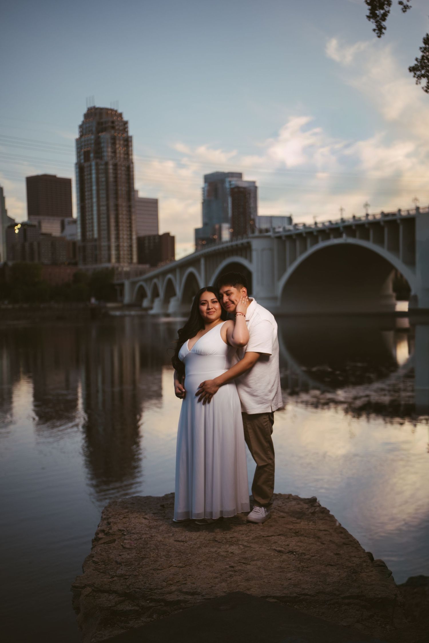 Couple standing on a rock by the river with Minneapolis skyline and bridge reflected in the water.