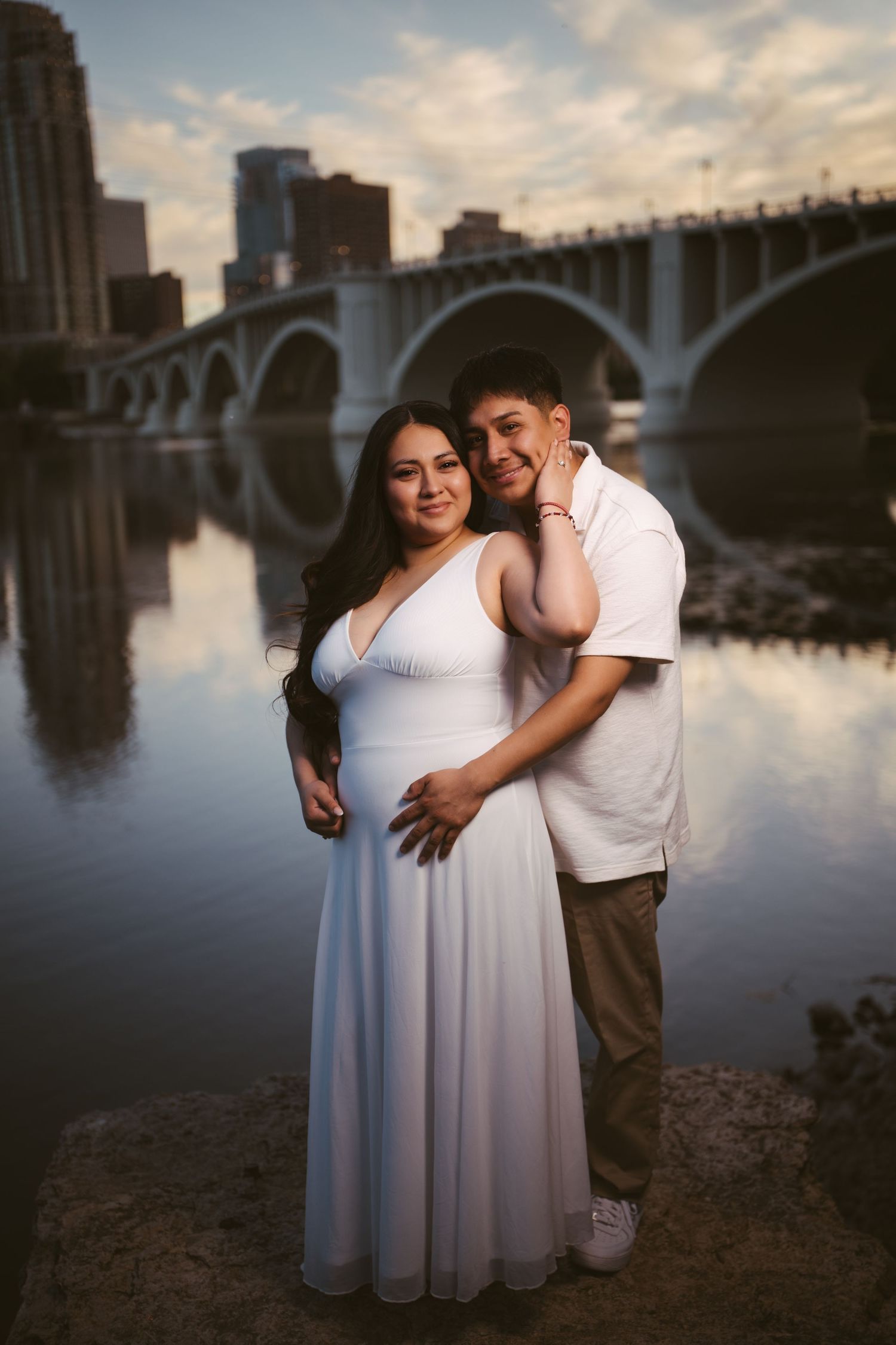Couple embracing near a river with a bridge and city skyline reflected in the water.