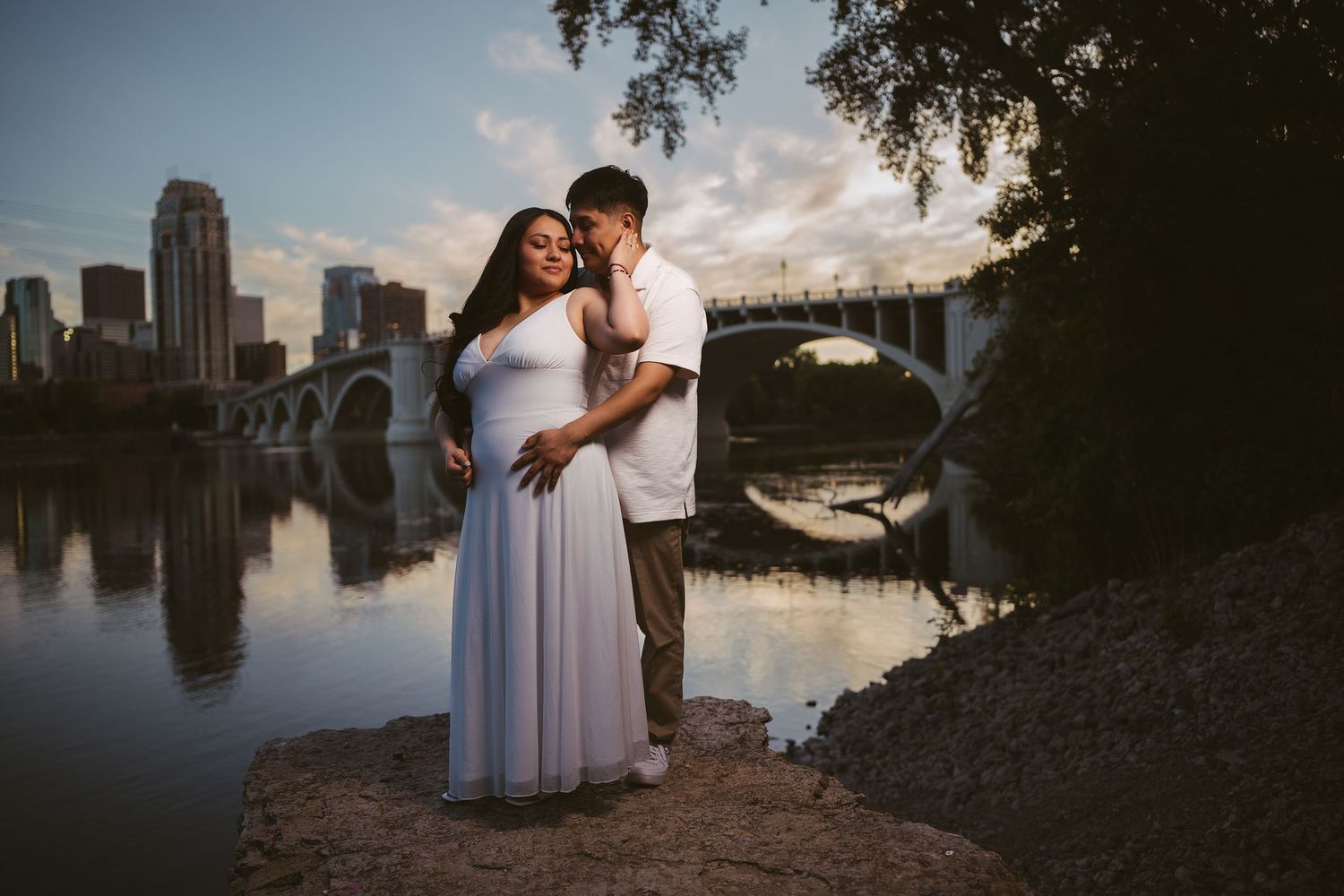 Couple embracing by a river with a city skyline and bridge reflecting in the water at golden hour.