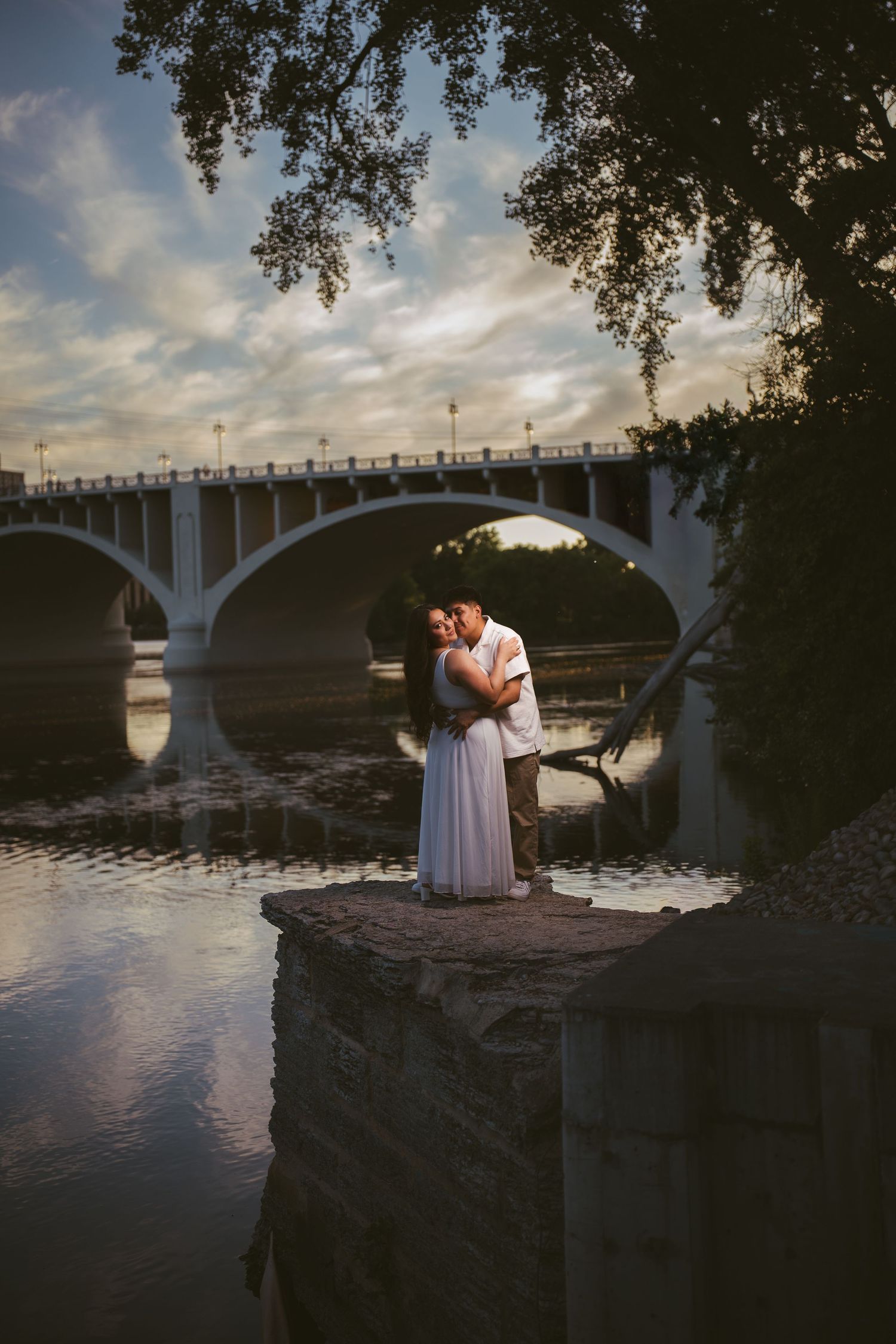 Couple embracing on stone pier at sunset with arched bridge and river reflection behind them.