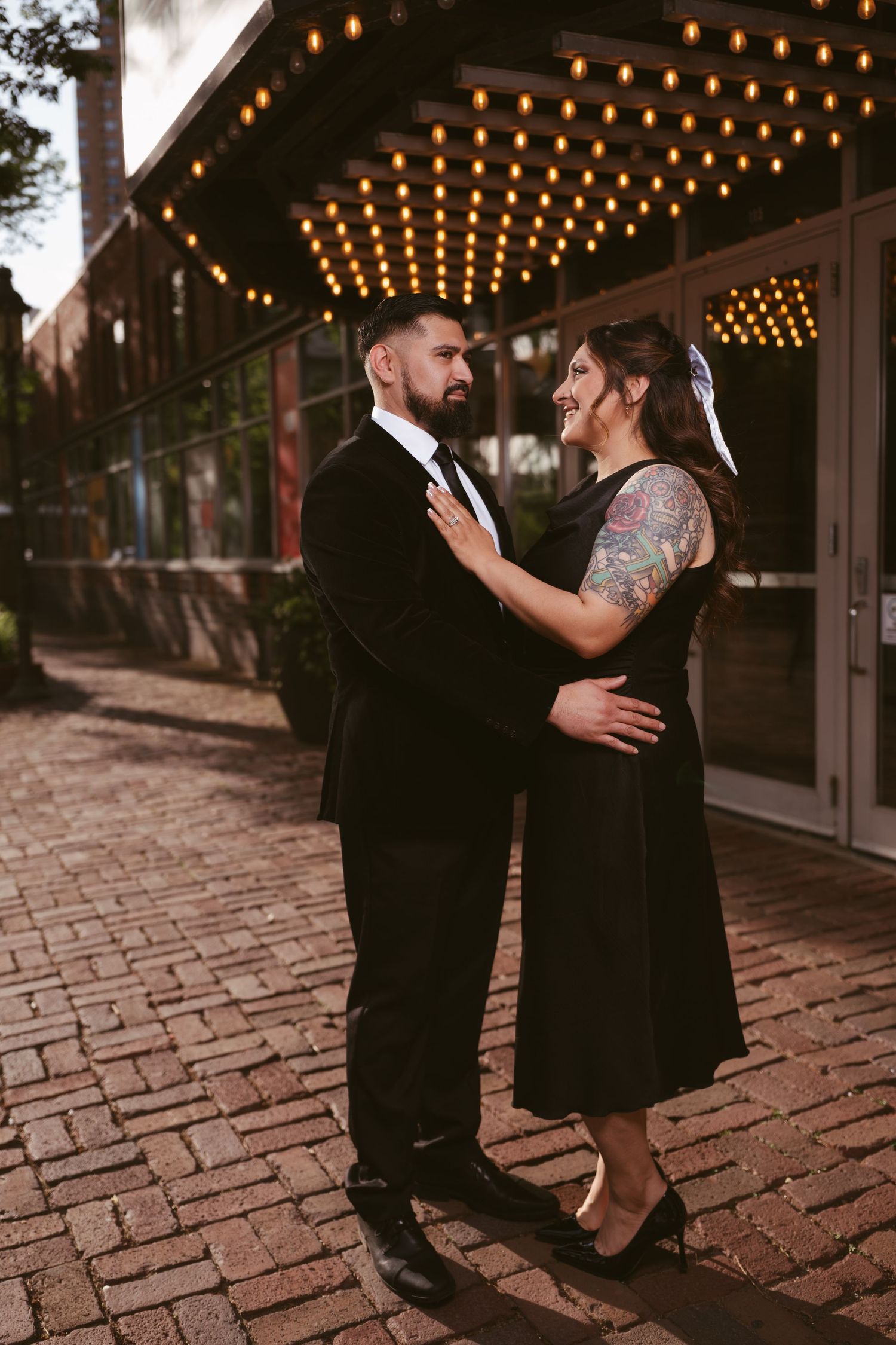 Couple in matching black outfits embracing under marquee lights on a brick sidewalk.