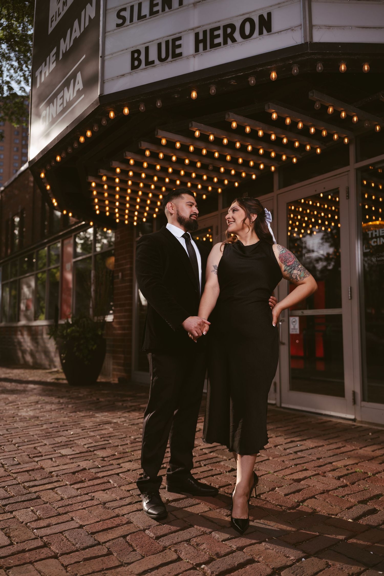 Couple in black attire holding hands outside The Main Cinema under marquee lights displaying Blue Heron on brick sidewalk.