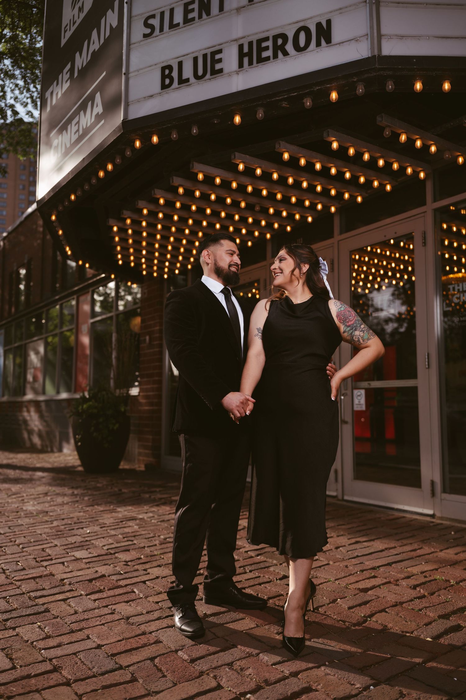 Couple in black attire holding hands outside Blue Heron Cinema marquee with glowing lights at dusk.