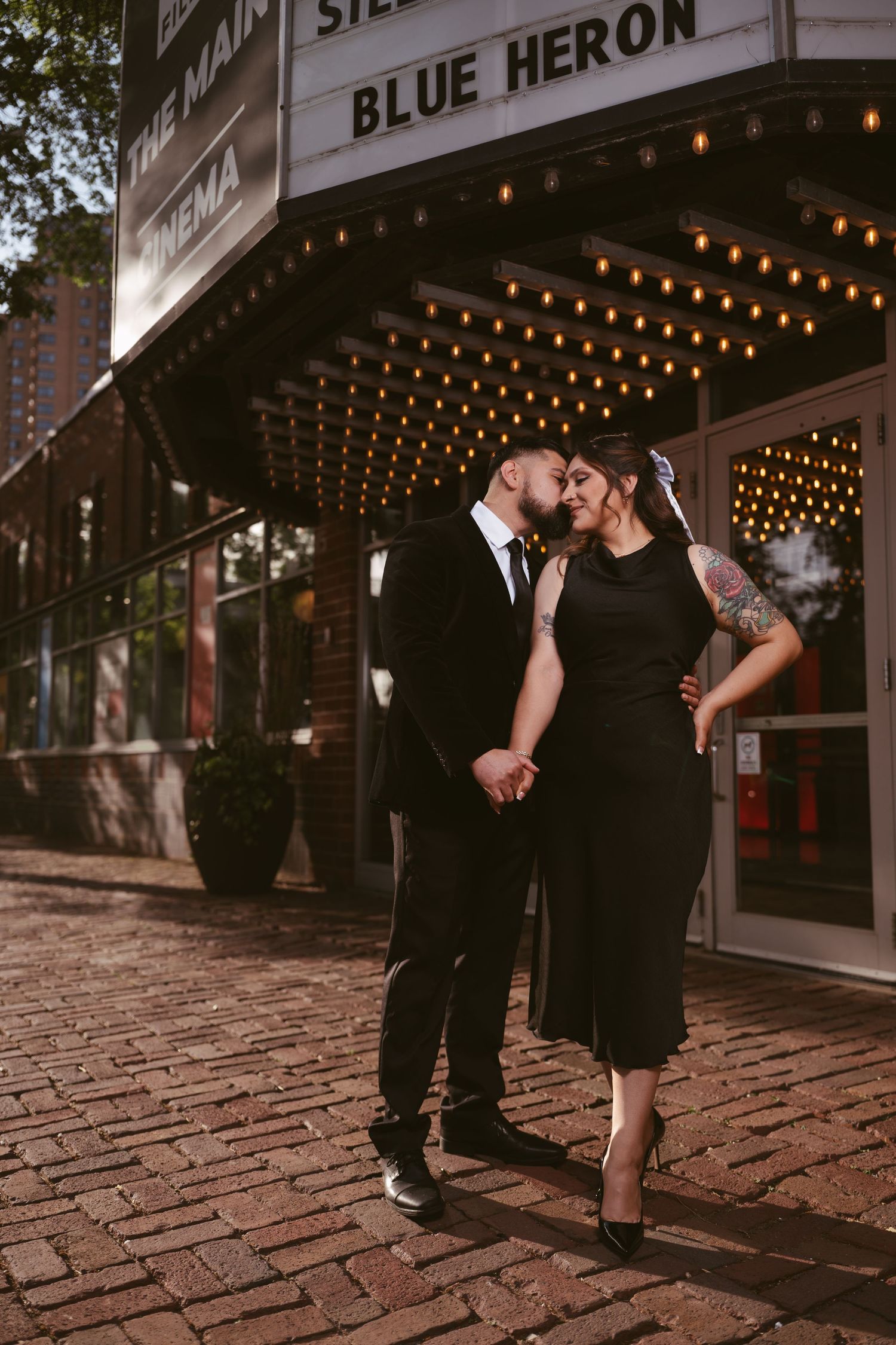 Couple in black attire sharing a romantic moment outside the Blue Heron Cinema marquee on a brick sidewalk.