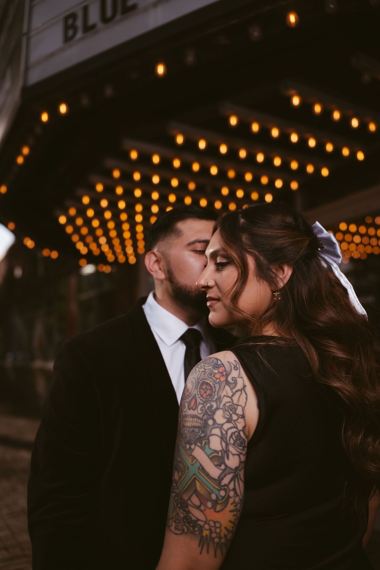 Couple in black formal wear share a romantic moment under glowing marquee lights outside the Blue theater at night.